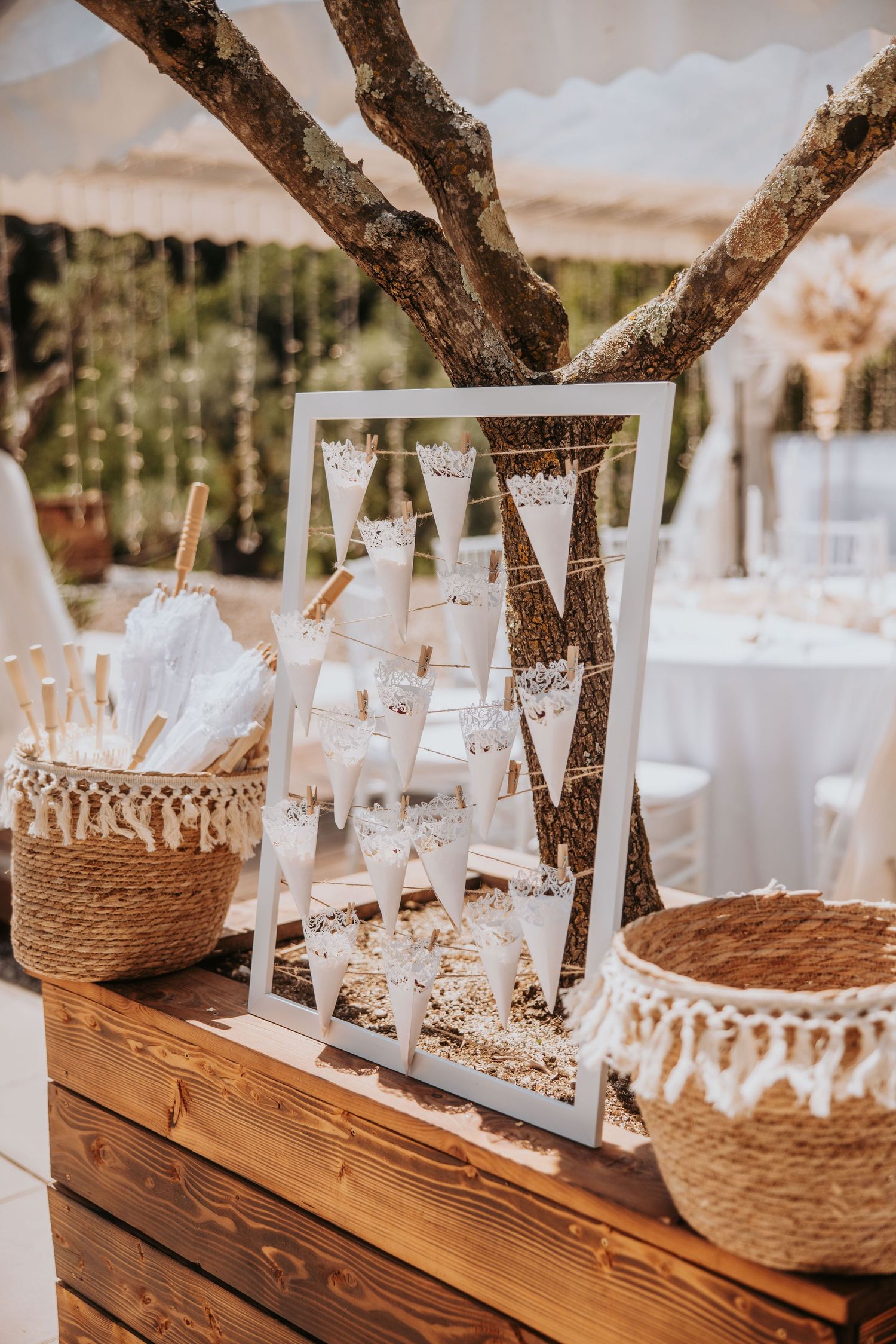 Rustic wedding dessert table with white frame display, lace bunting, and wicker baskets under a tree.