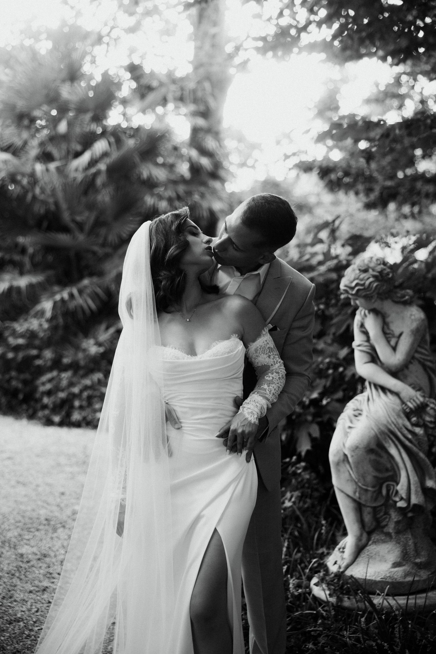 Black and white romantic kiss between bride and groom near garden statue.