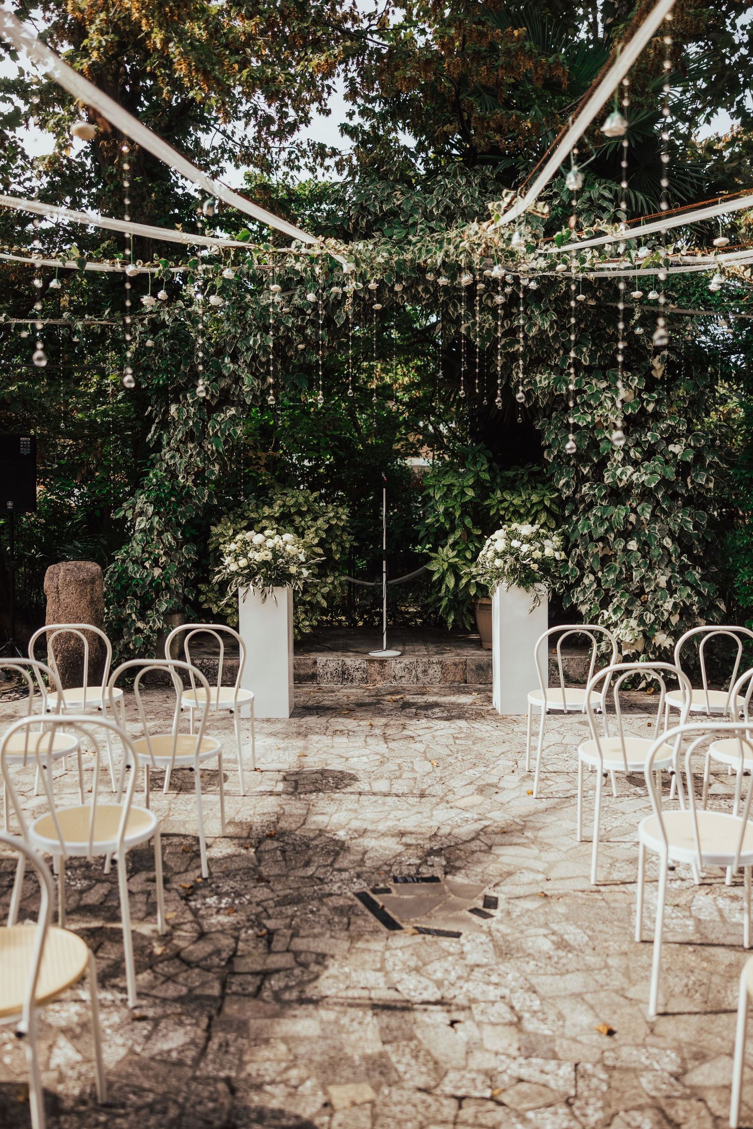 Elegant outdoor wedding ceremony setup with white chairs, floral arrangements, and hanging crystal decorations.