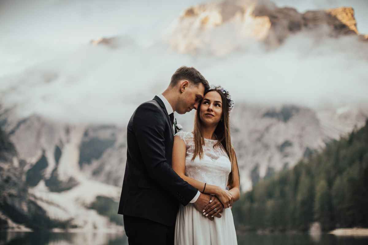 Romantic wedding couple embracing by a serene mountain lake with misty Dolomites peaks behind them.
