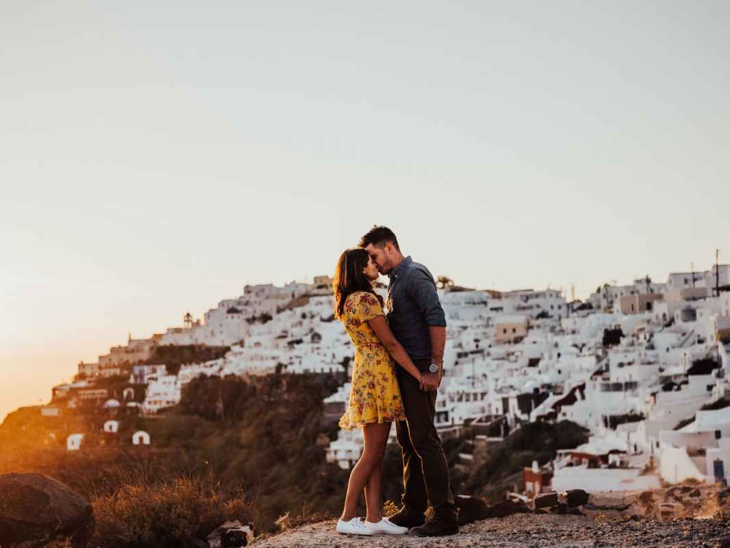 Couple sharing a romantic kiss at sunset with white Santorini buildings on hillside backdrop.
