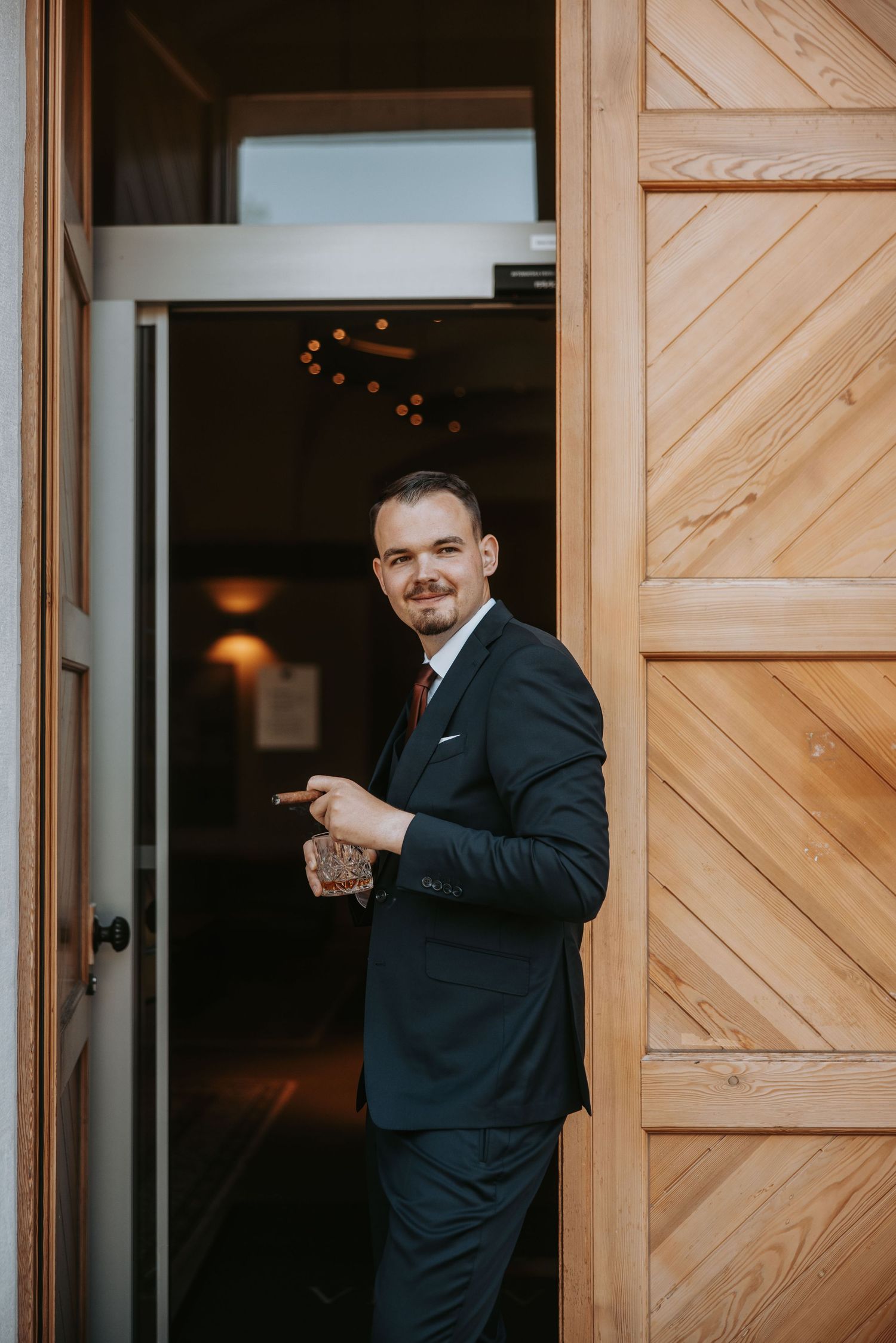 Dapper groom in navy suit smiling while holding a gift, standing at a wooden barn door entrance.