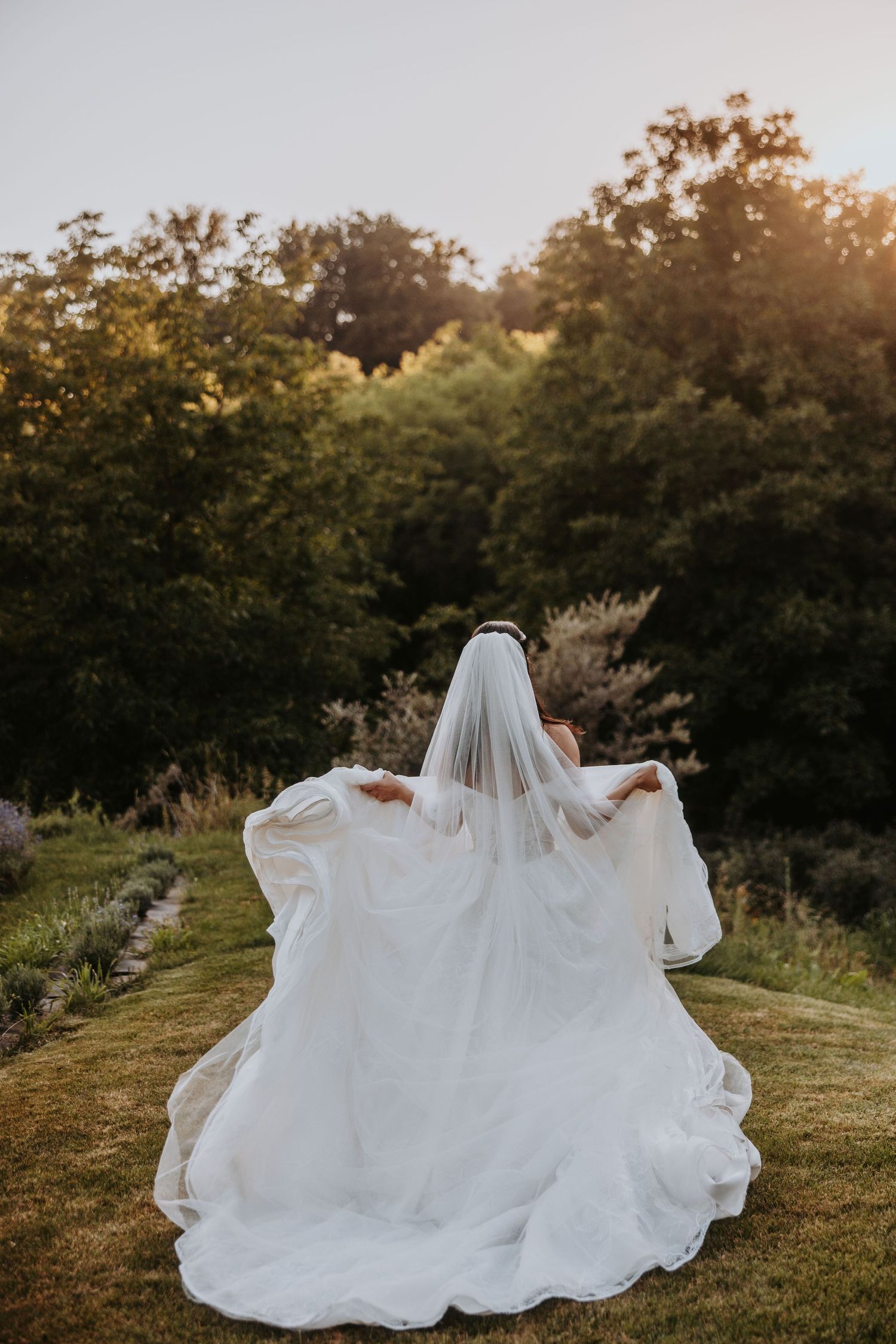 Bride in a flowing white gown and veil walks through a lush garden at golden hour sunset.