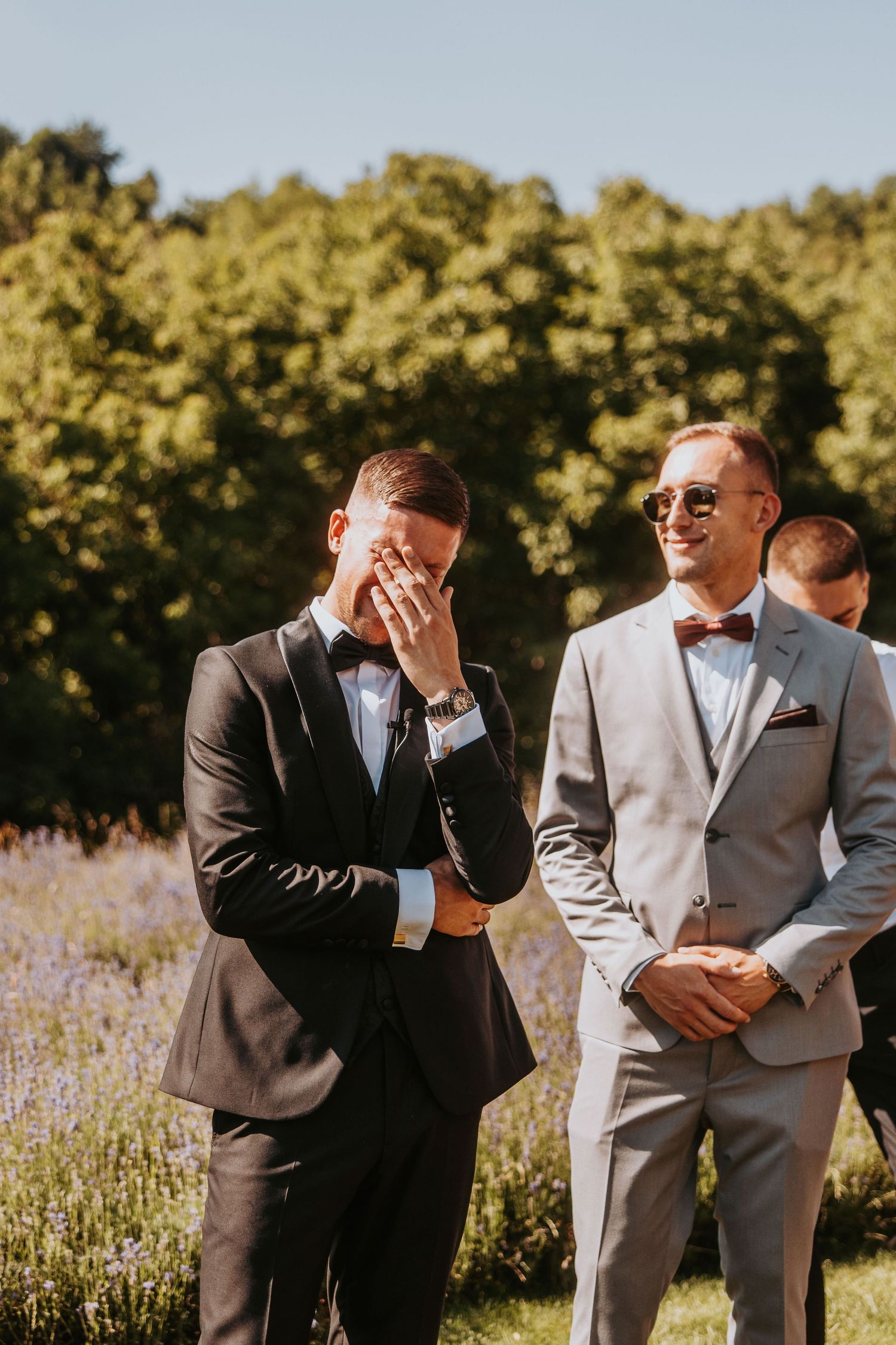 Emotional groom wiping tears at outdoor wedding ceremony beside smiling best man in gray suit and bow tie.