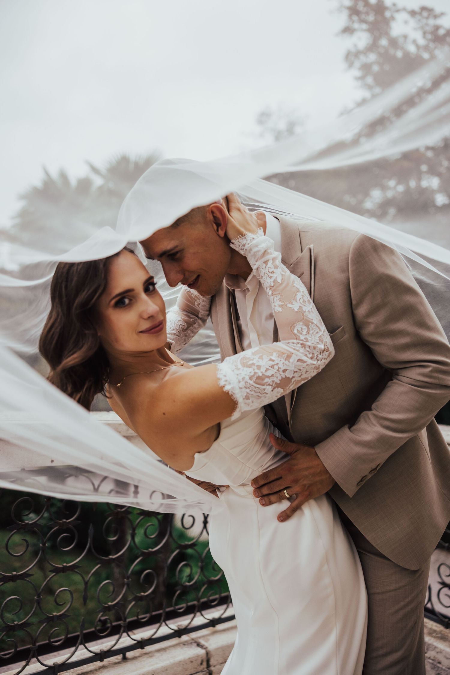 Bride and groom share romantic moment under flowing white veil, groom wearing tan suit and white cowboy hat.