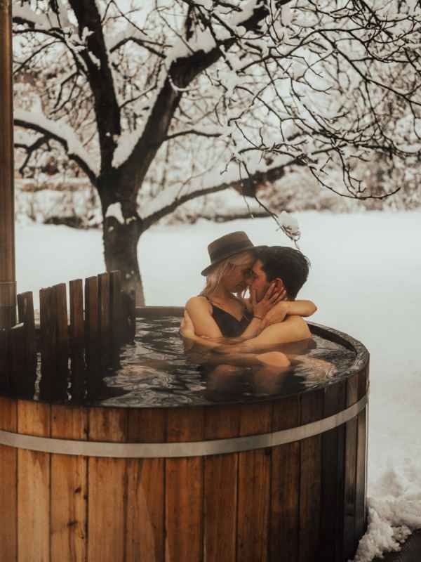 Couple kissing in a wooden hot tub surrounded by snow-covered trees in a romantic winter outdoor setting.