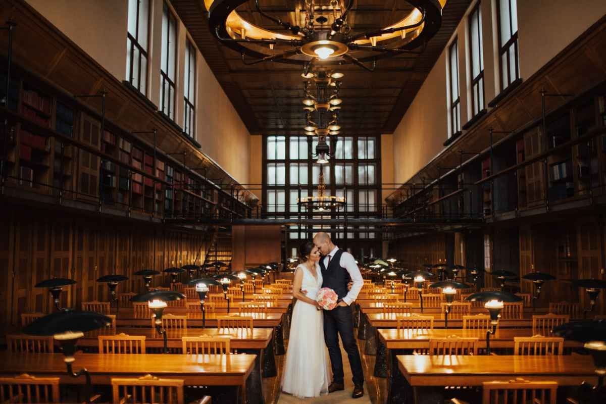 Bride and groom embrace in a grand library wedding venue with warm lighting and tall bookshelves.