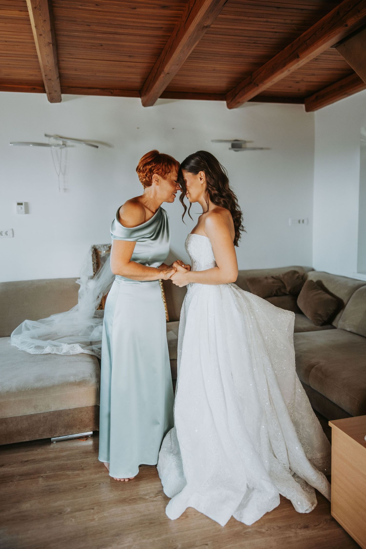 Bride and bridesmaid in elegant gowns share an intimate moment holding hands in a bright wedding preparation room.