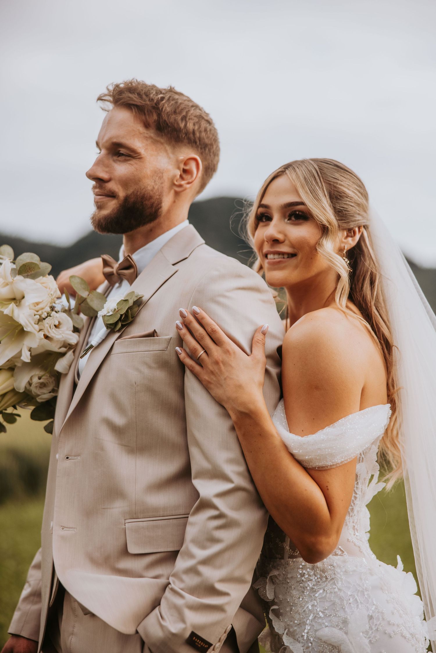 Smiling bride in lace dress stands beside groom in beige suit holding white bouquet outdoors.