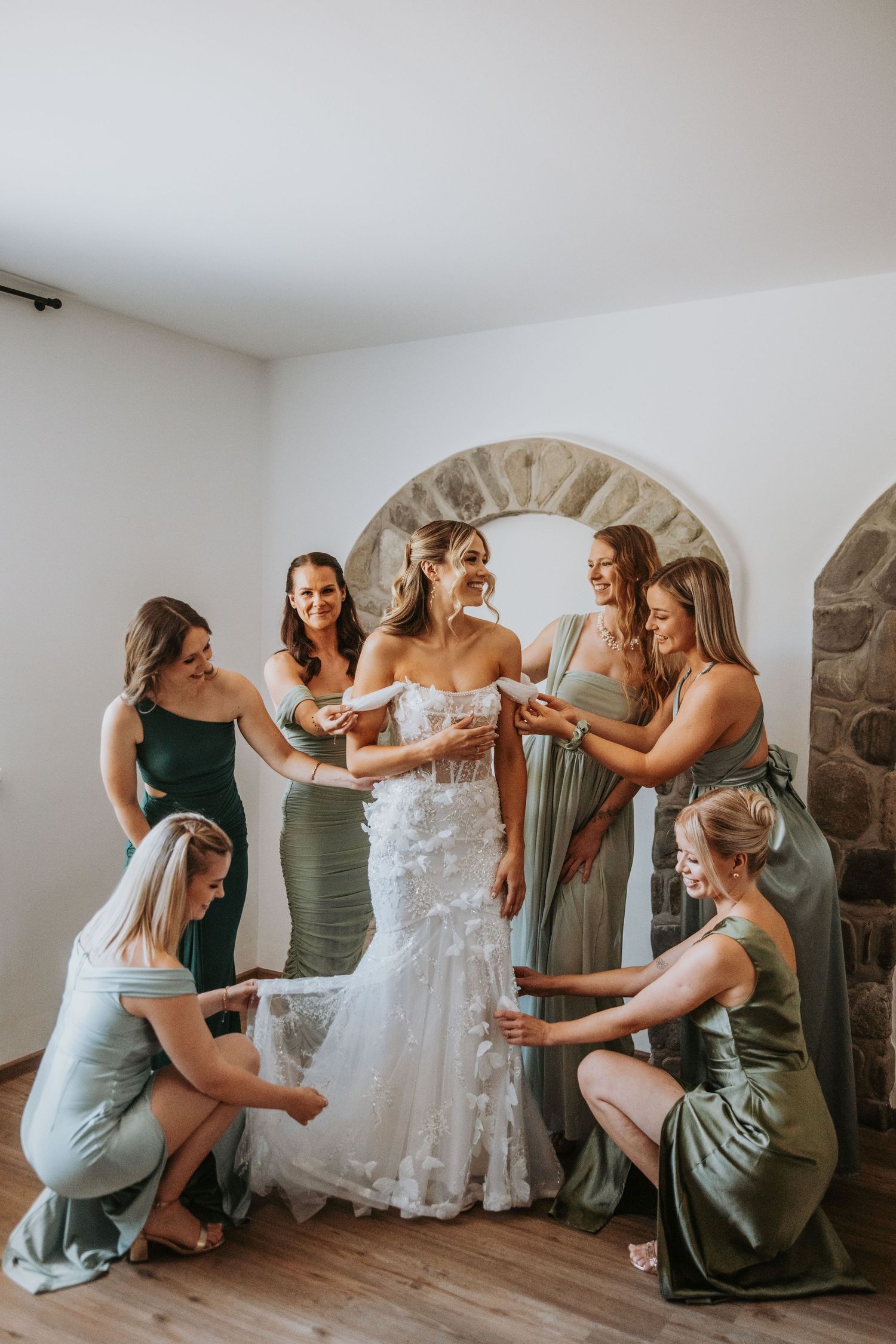 Bride in strapless lace gown surrounded by six bridesmaids in sage green dresses during wedding preparation.