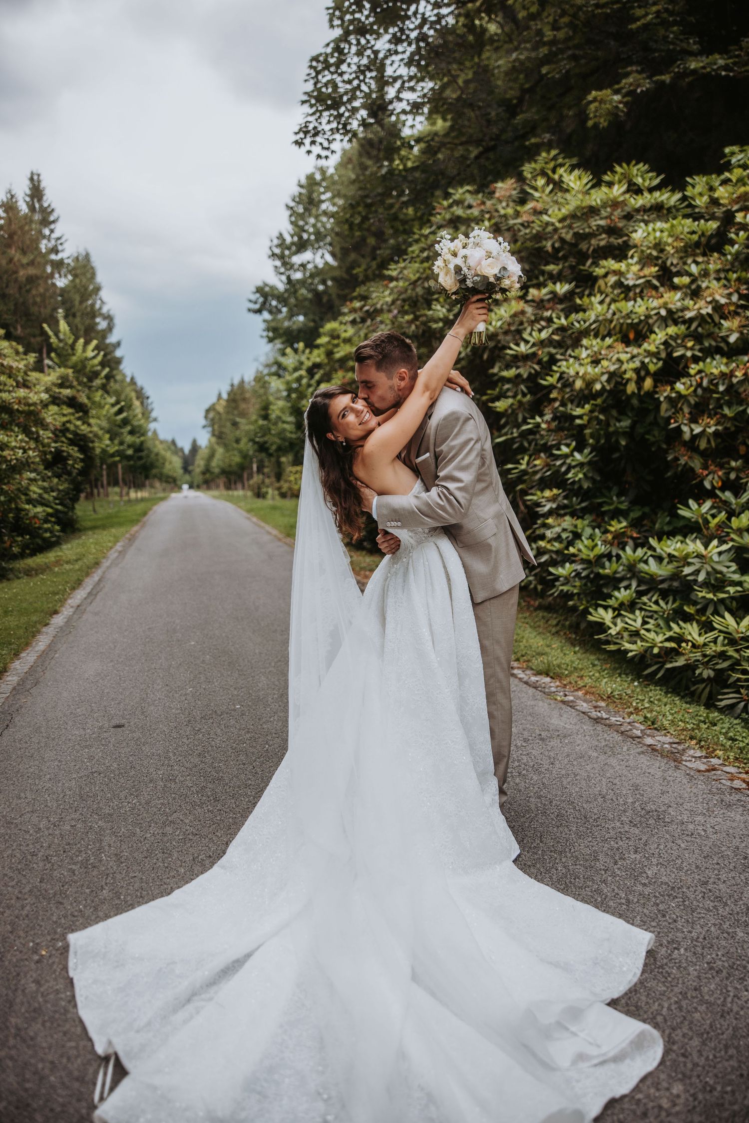 Bride and groom embracing on tree-lined road, bride in flowing white gown with long veil, groom in tan suit.
