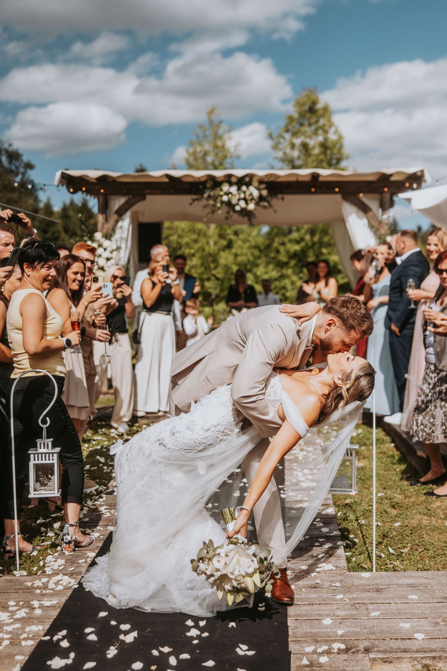 Bride and groom share a romantic dip kiss at outdoor wedding ceremony while guests celebrate with confetti.