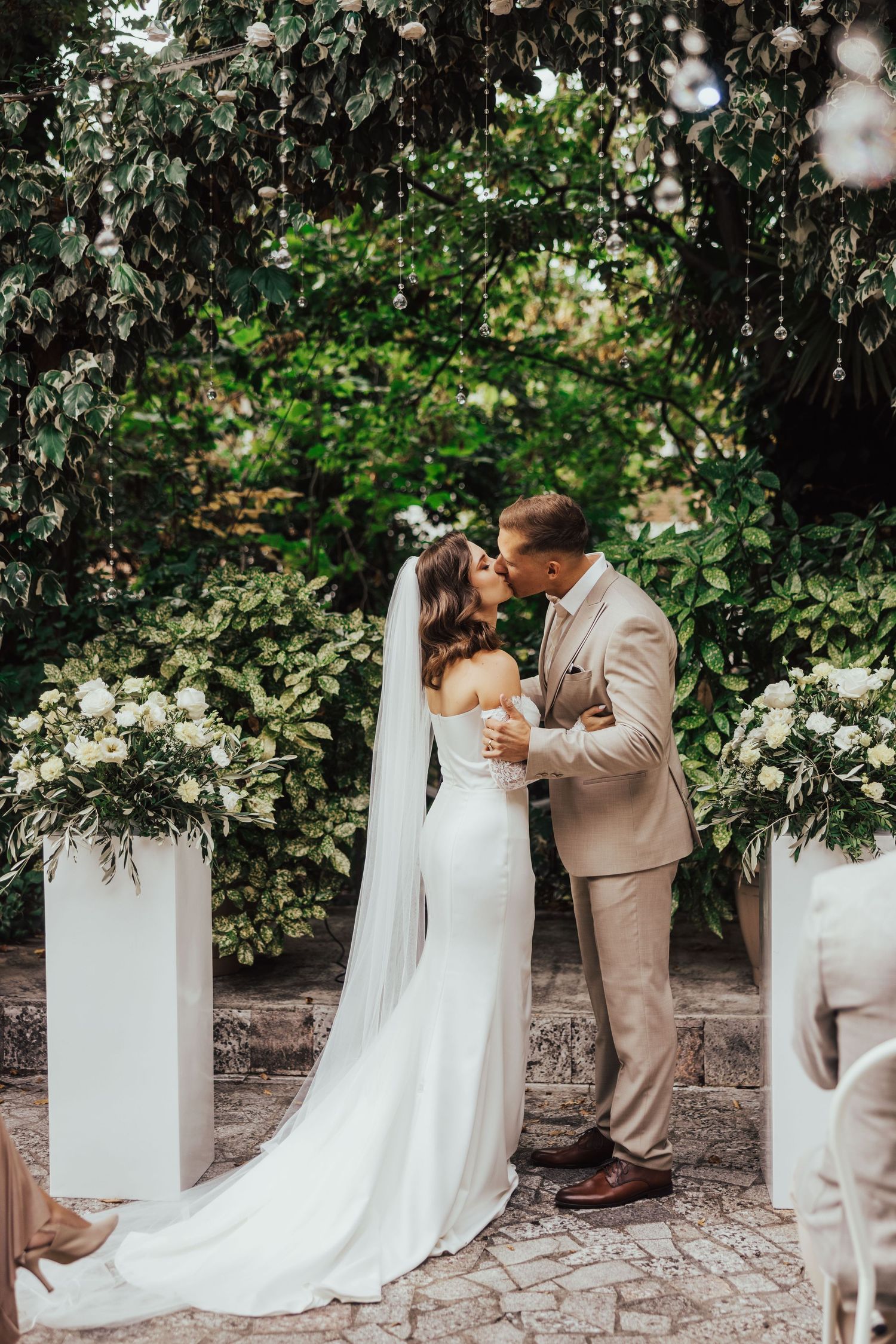 Newlyweds share first kiss at elegant outdoor wedding ceremony with white flowers and green foliage backdrop.