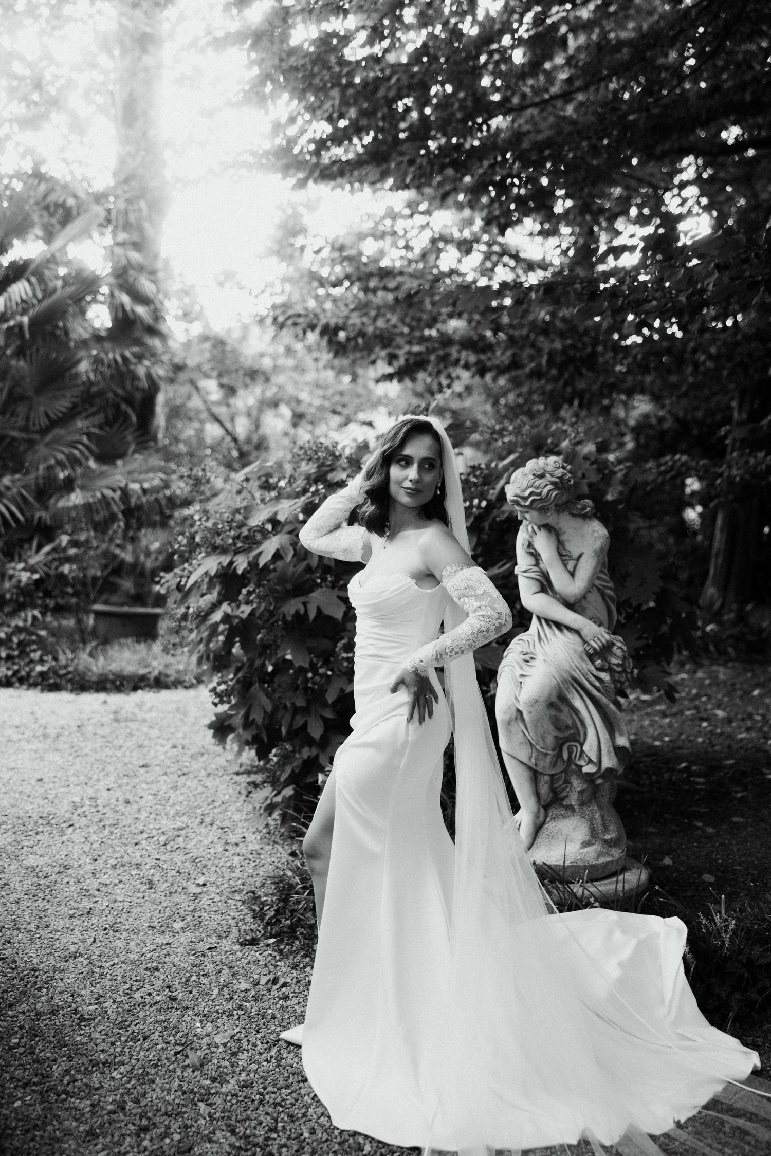Elegant bride in white gown and veil poses beside stone angel sculpture in a lush garden setting.