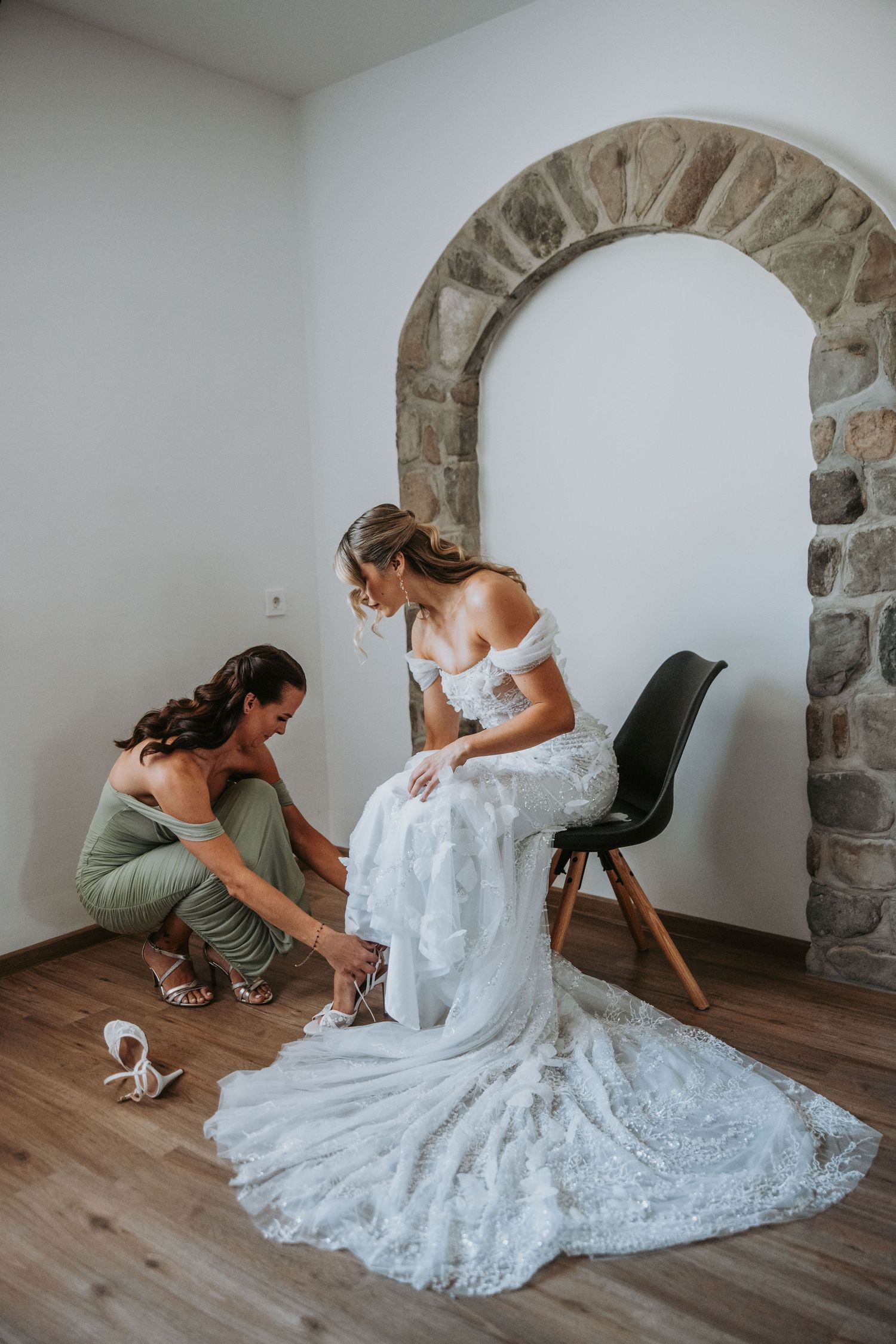 Bridesmaid helping bride put on heels while seated on chair in room with stone arch decor.