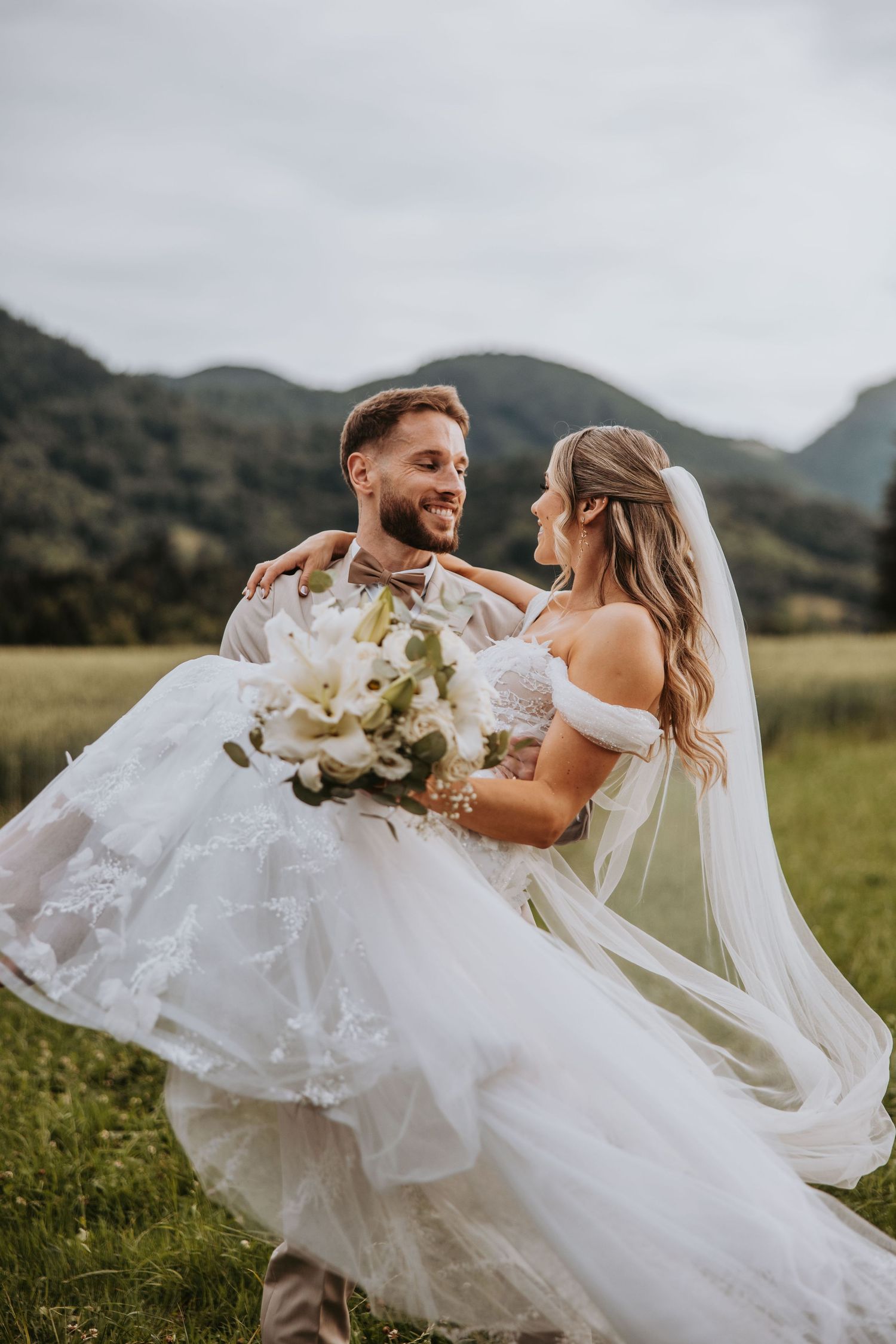 Joyful groom lifts smiling bride in lace wedding gown in a green meadow with mountain scenery.