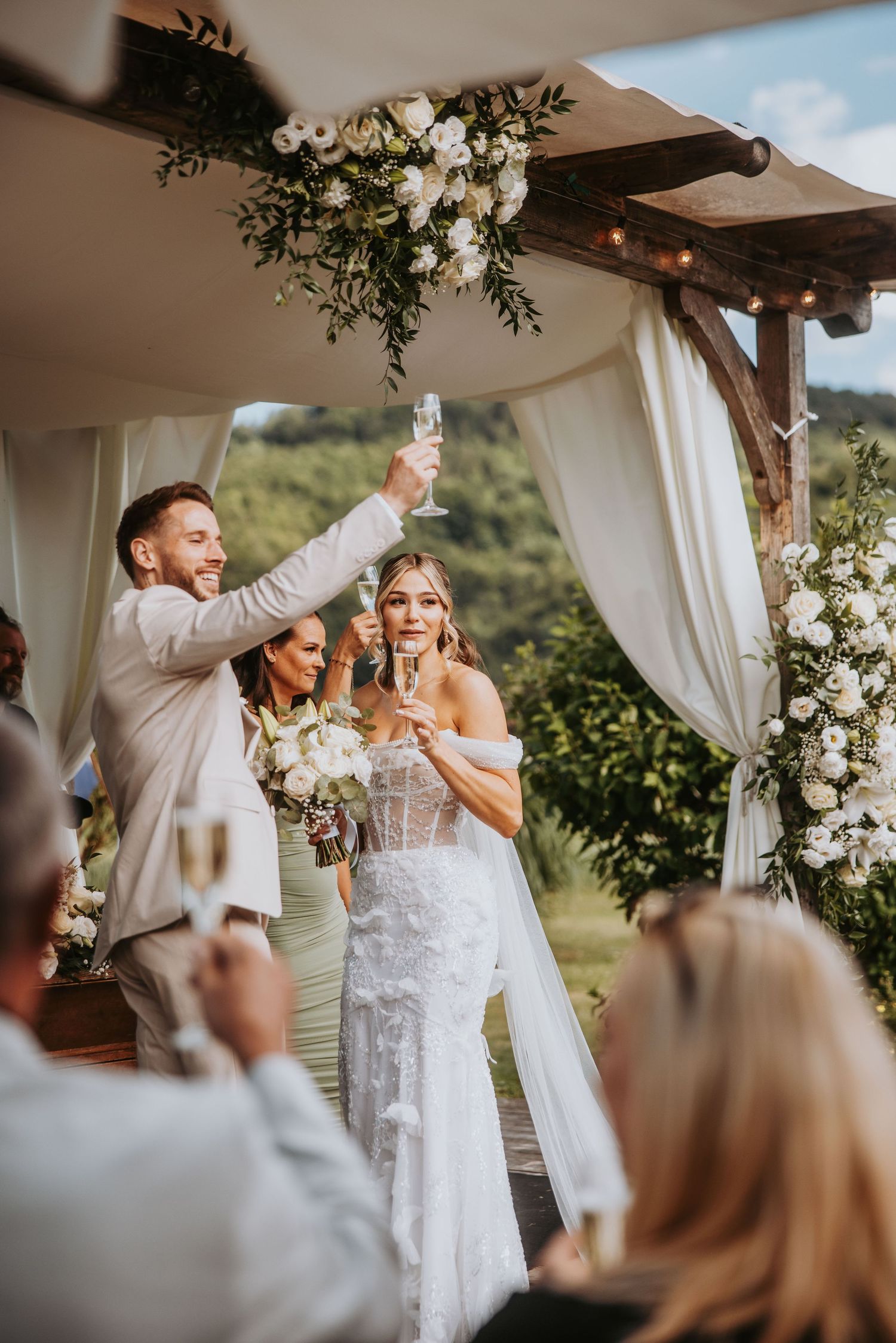 Bride and groom raise champagne glasses in toast under floral pergola at outdoor wedding reception.