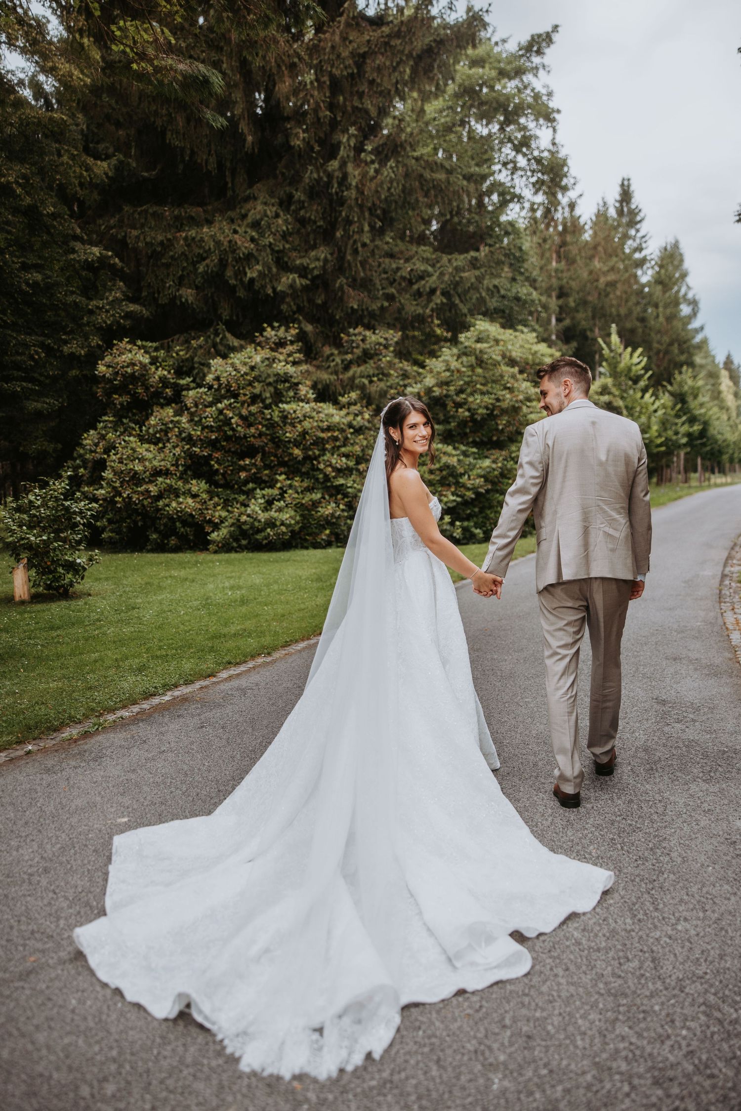 Bride and groom walk hand-in-hand on a tree-lined path, her cathedral veil trailing elegantly behind.