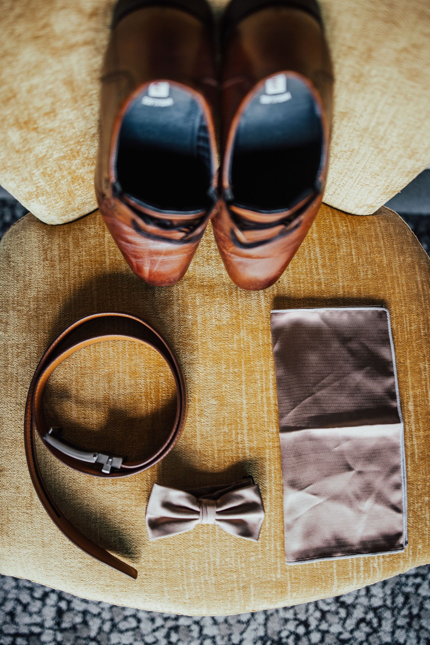 Grooms wedding accessories flat lay featuring brown leather shoes, belt, bow tie, and pocket square on chair.