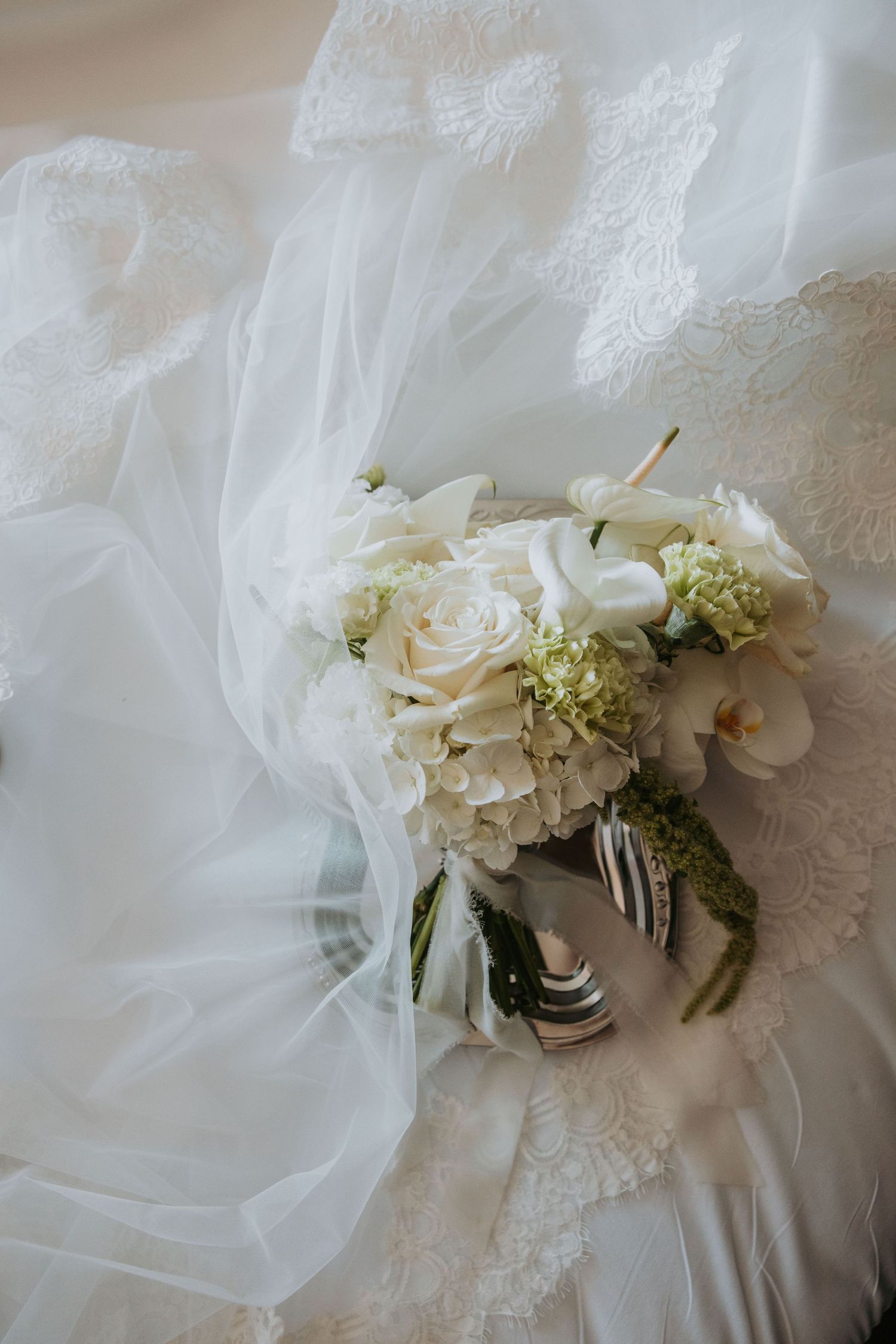 Romantic bridal bouquet of white roses, hydrangeas, and orchids resting on lace wedding dress with tulle veil.