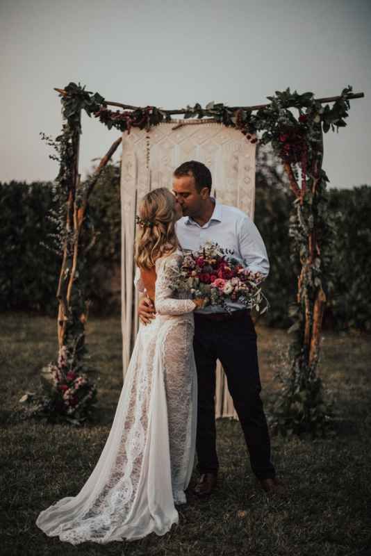 Bride and groom share first kiss under rustic floral arch with burgundy blooms at outdoor wedding.