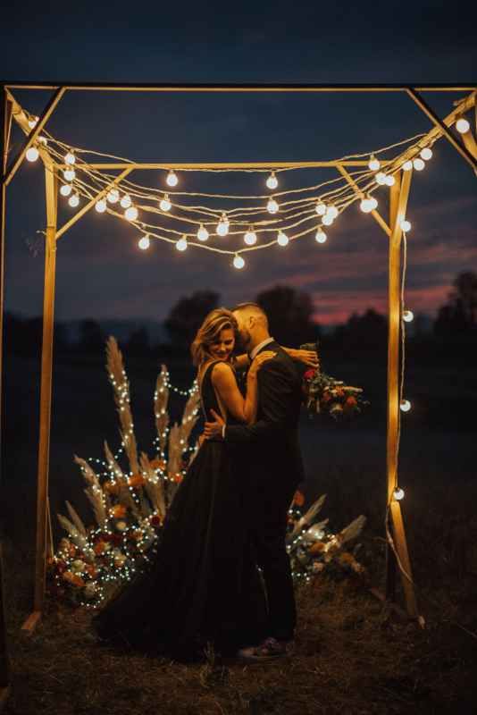 Newlyweds embracing under glowing string lights wedding arch at twilight with floral arrangements.