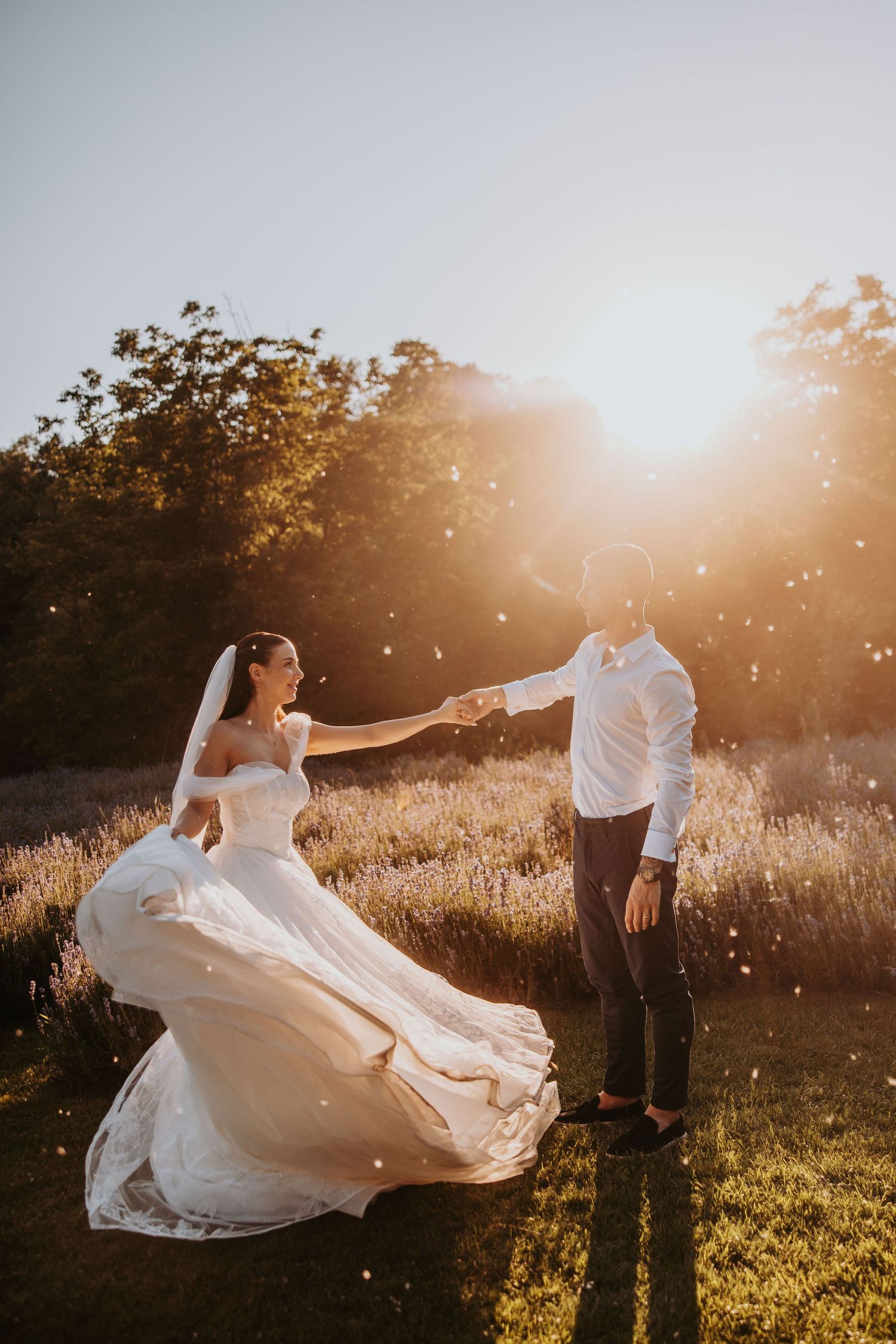 Bride and groom dancing in golden sunset light in lavender field, bride's gown flowing as they hold hands.