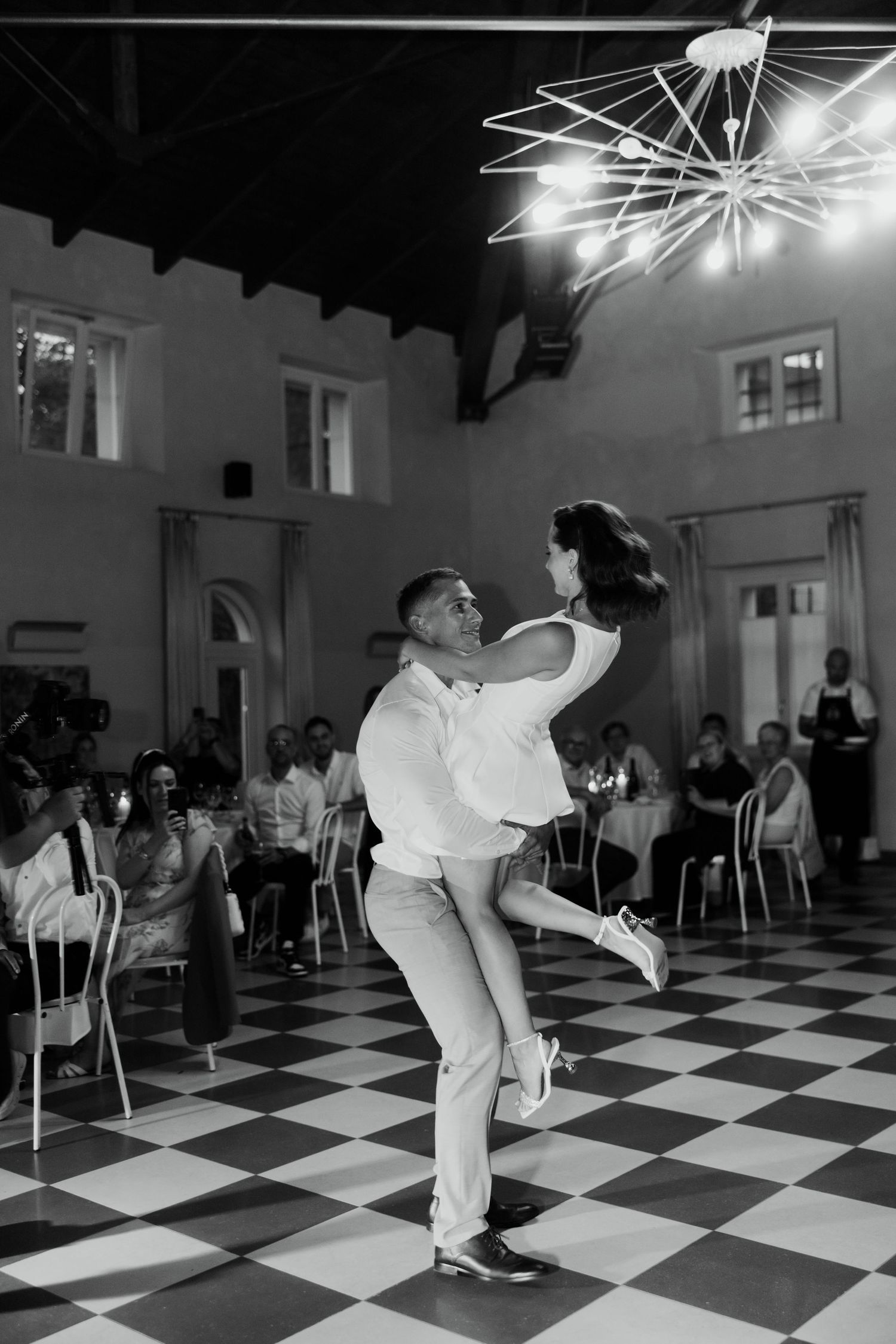 Bride and groom sharing energetic first dance on checkered floor at wedding reception.