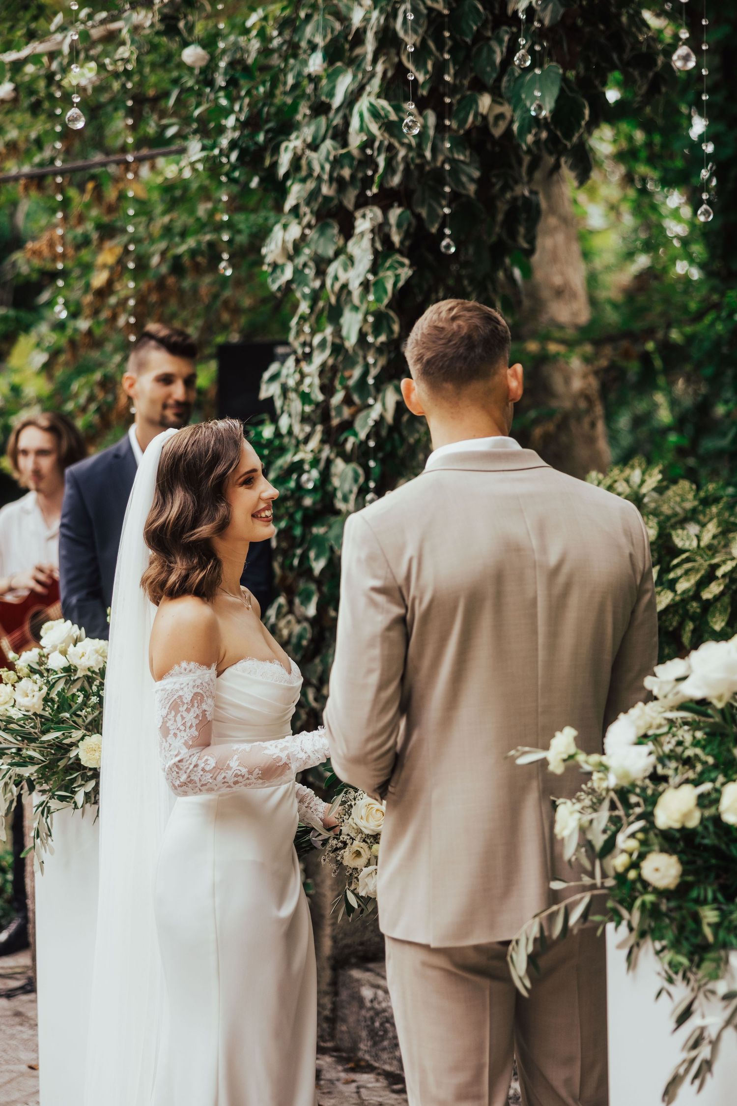 Smiling bride in lace wedding dress exchanging vows with groom at outdoor ceremony.