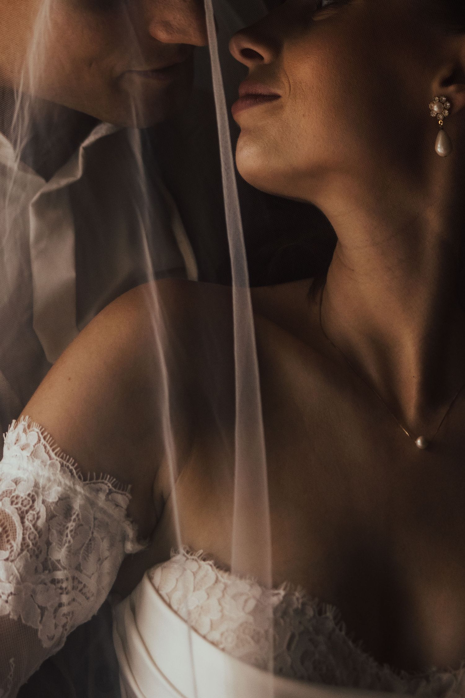 Bride in lace off-shoulder gown with pearl earrings and necklace, draped under a sheer white wedding veil.