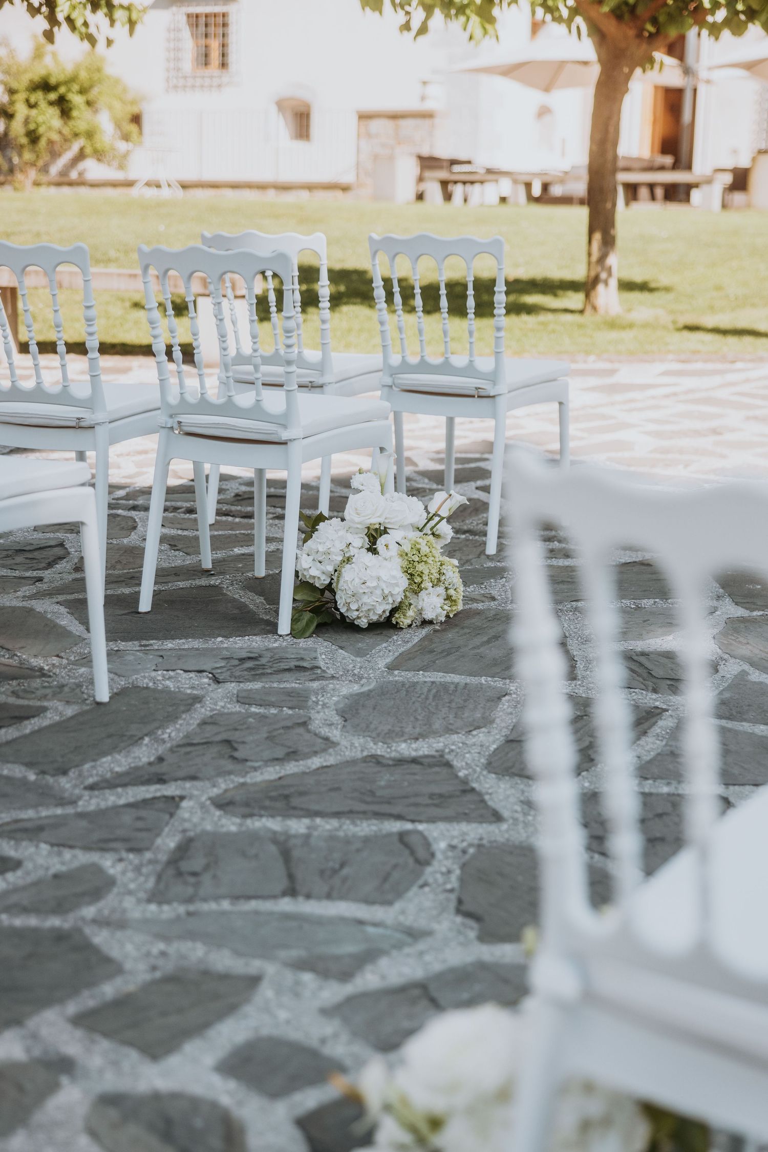 Elegant white Napoleon chairs arranged on stone patio with hydrangea bouquets for outdoor wedding ceremony.