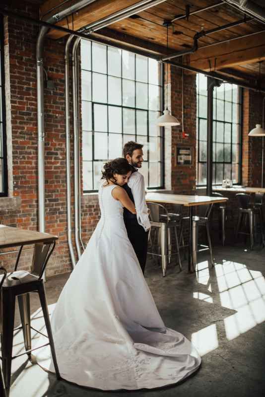 Bride and groom embracing in a sunlit industrial brick venue with large factory windows.