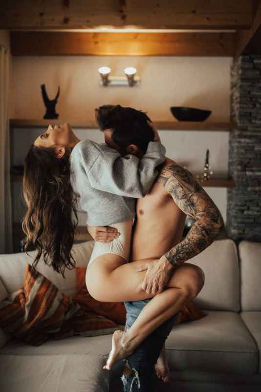 Tattooed man and woman in an intimate embrace on a couch in a cozy rustic living room.