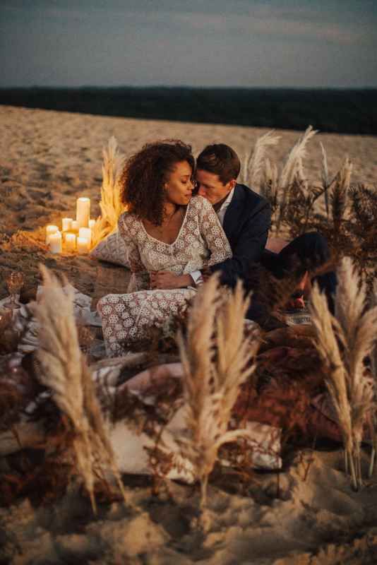 Couple sharing an intimate moment on a sandy beach surrounded by candles and pampas grass at dusk.