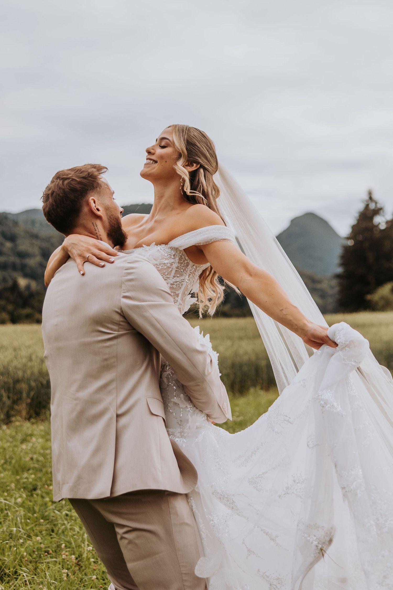 Groom lifting joyful bride in lace wedding gown in green field with mountains.