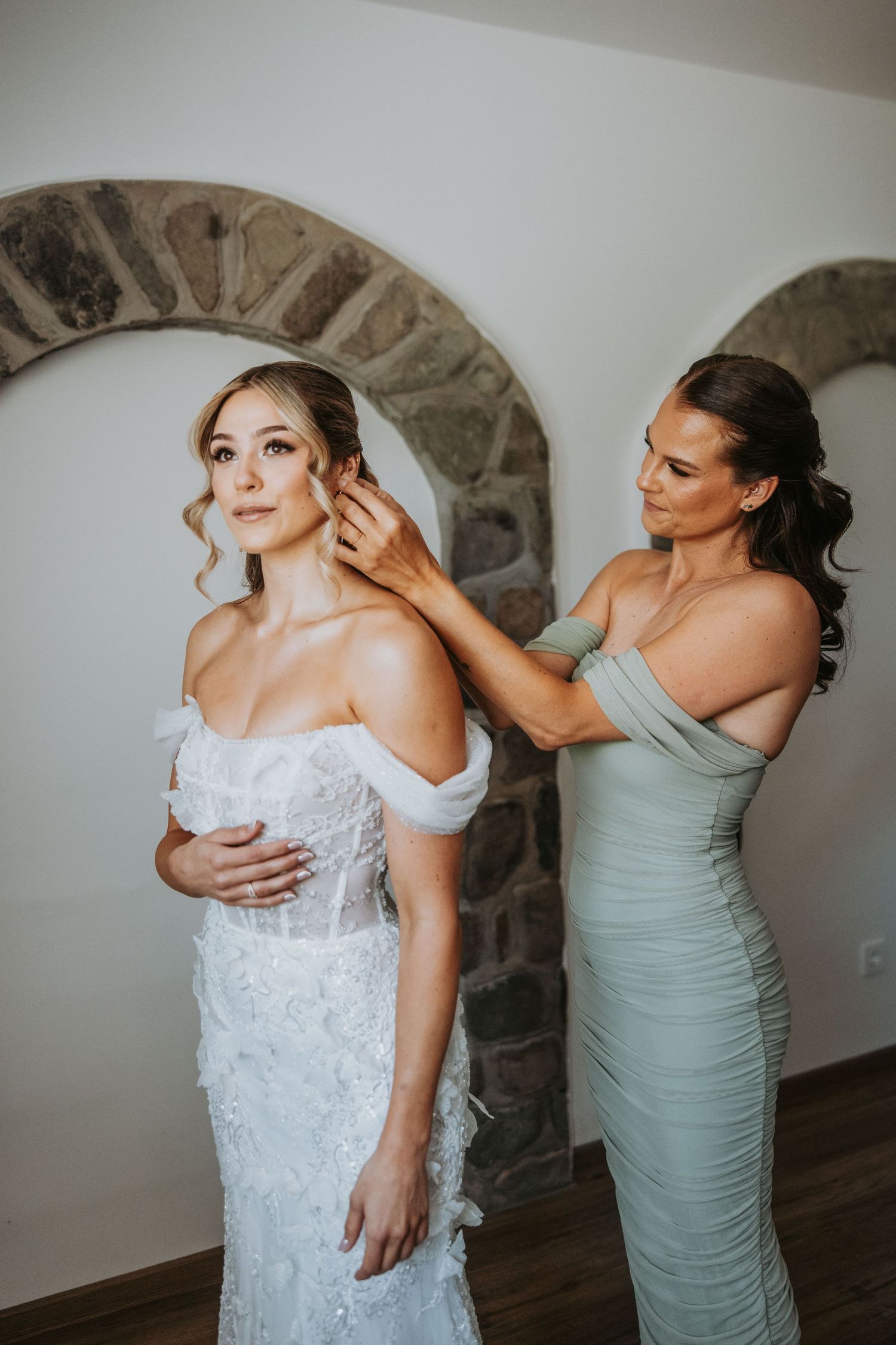 Bridesmaid adjusting bride's earring as she stands in elegant off-shoulder lace wedding gown.