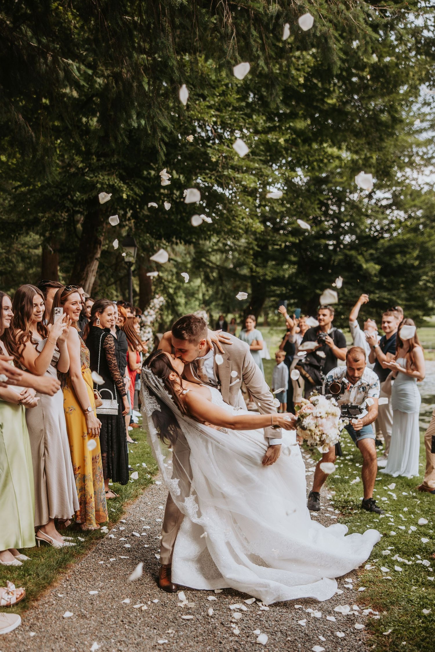 Groom dips bride for a romantic kiss as wedding guests toss white flower petals along an outdoor aisle.