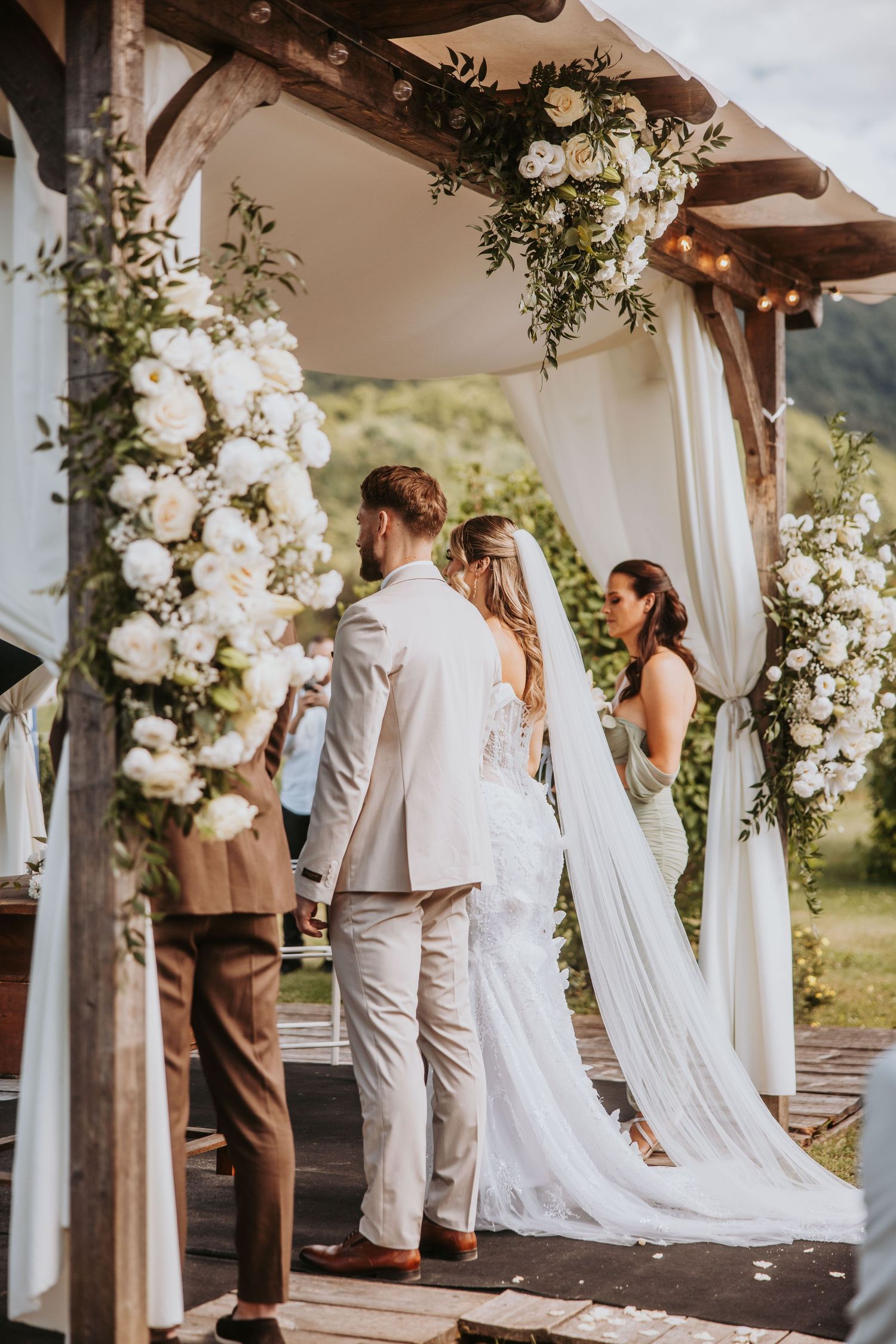 Bride and groom exchanging vows under floral arch adorned with white roses and greenery at outdoor wedding.