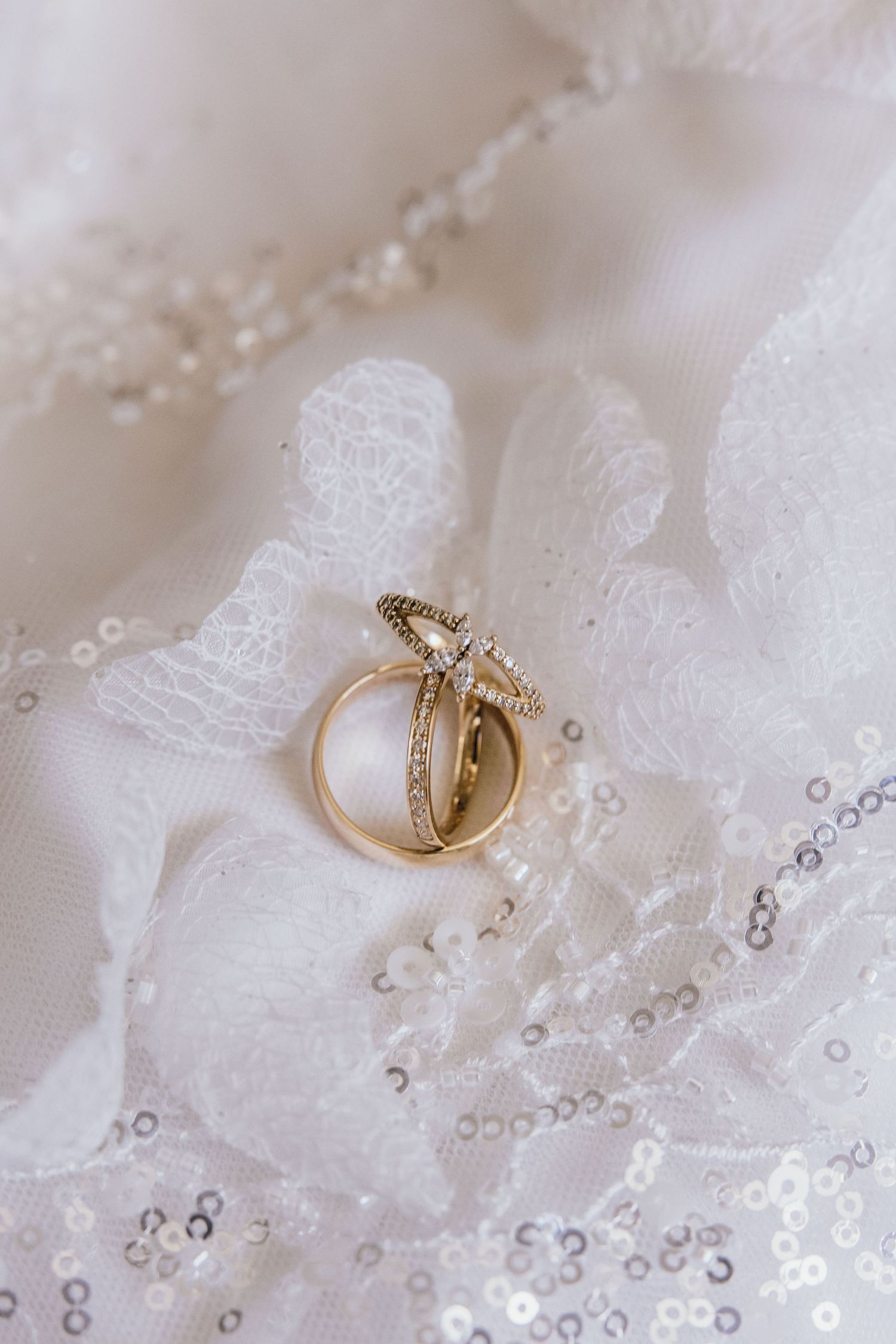 Gold wedding rings with diamond accents resting on white lace bridal fabric with sequin details.