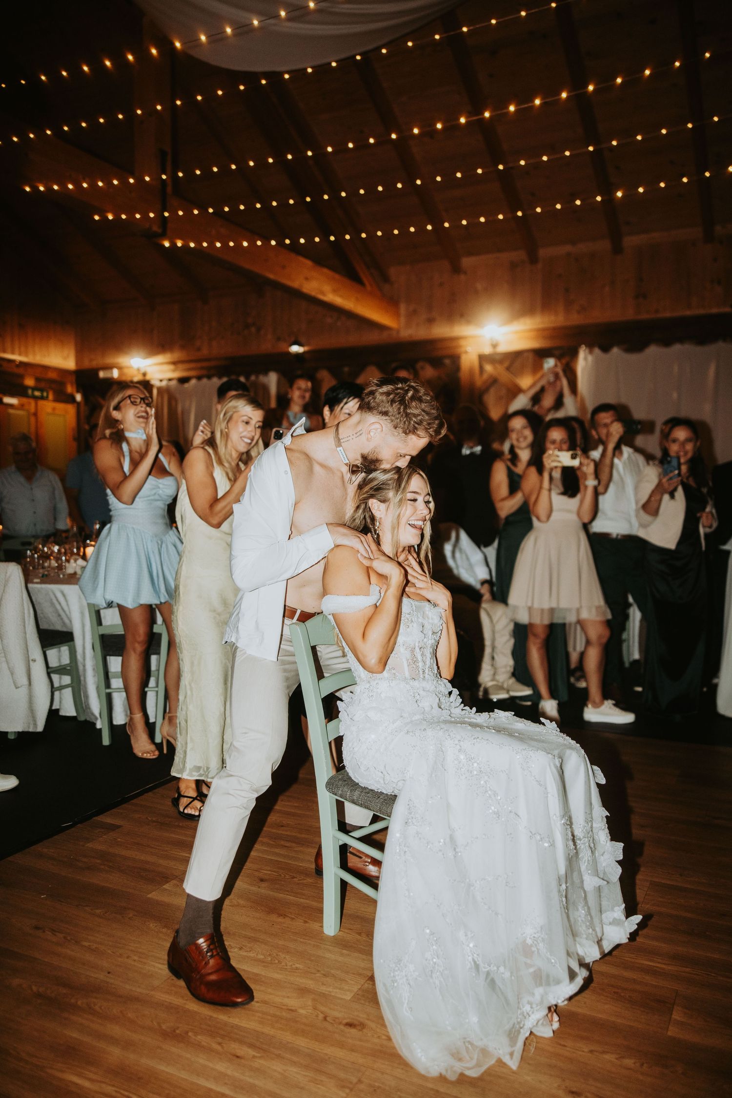 Groom kisses seated bride during lively wedding reception dance in rustic barn with string lights.