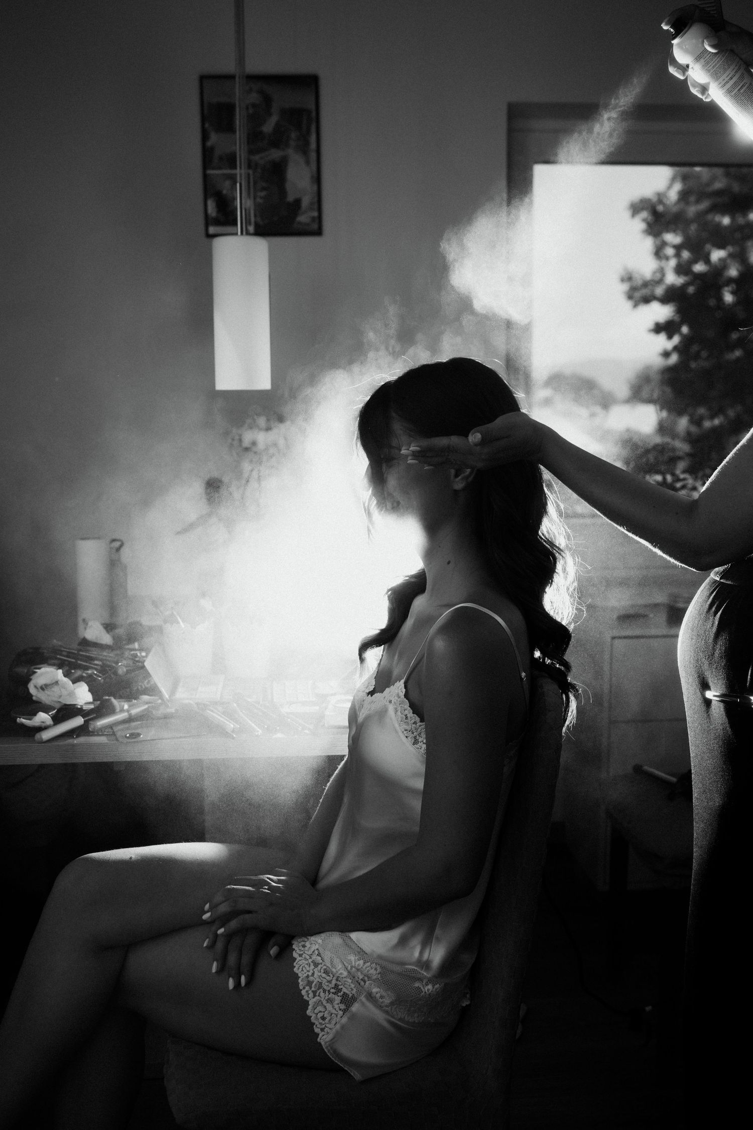 Black and white photo of a bride getting hair styled with hairspray mist in a softly lit dressing room.