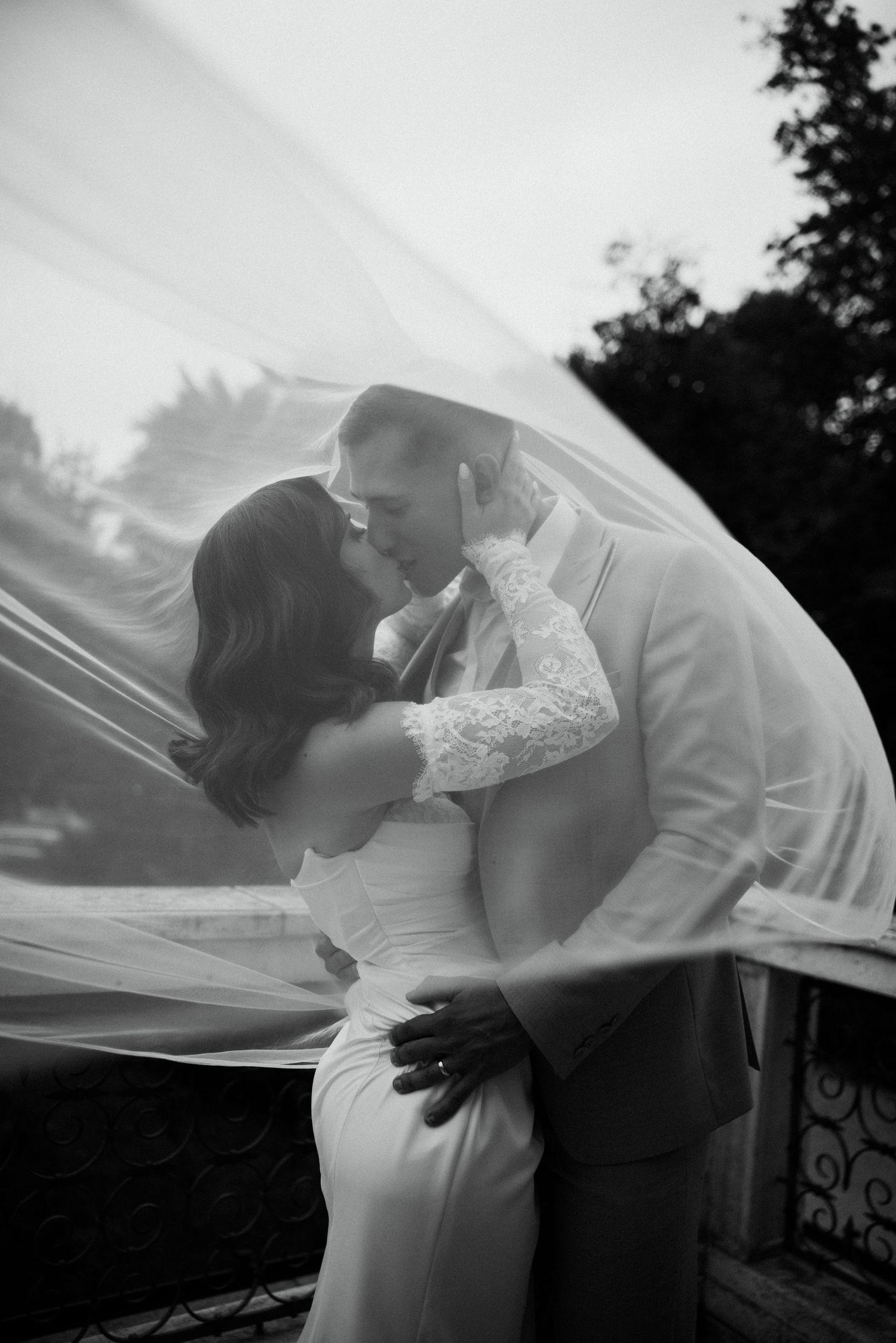 Black and white photo of bride and groom kissing under flowing wedding veil outdoors.