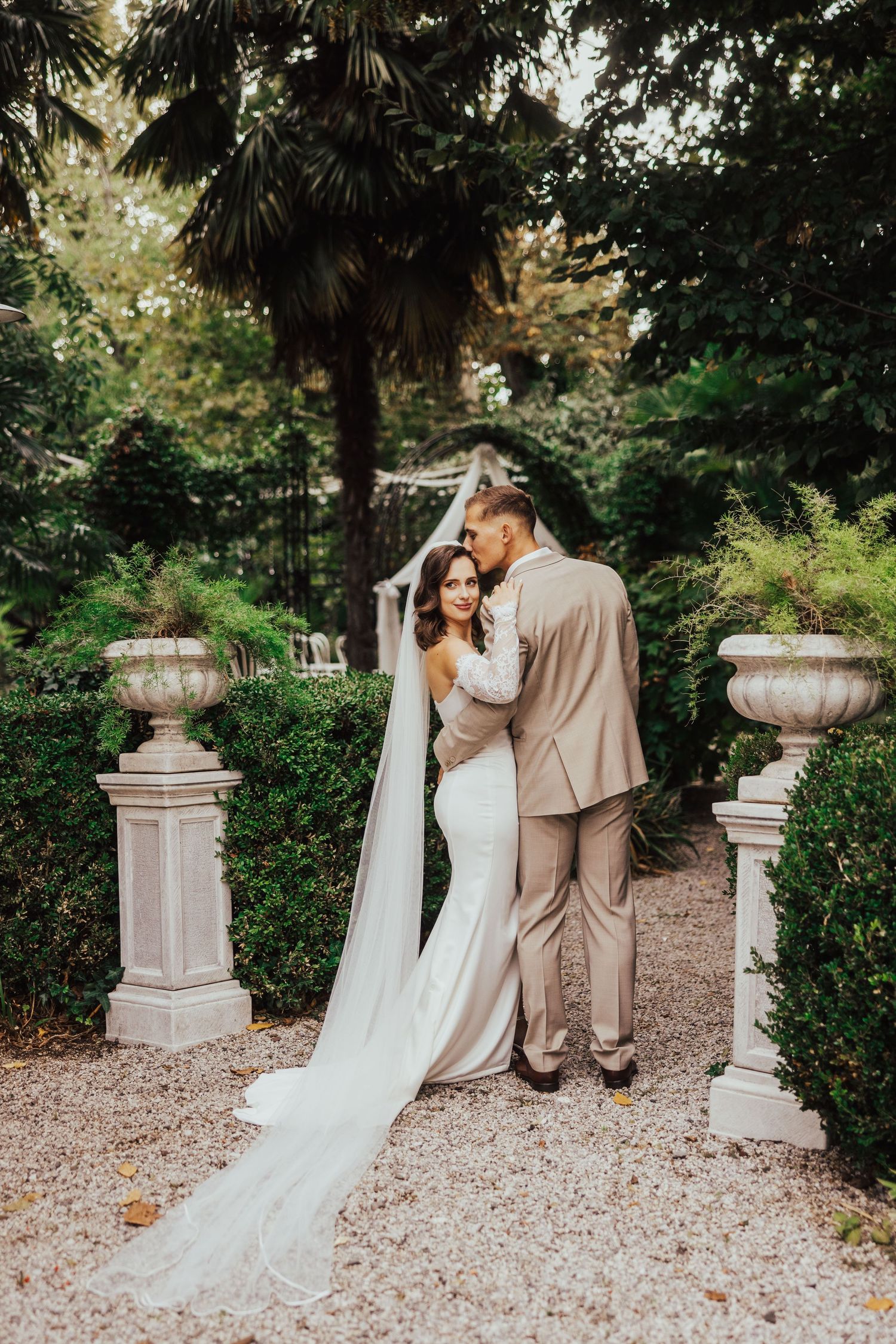 A bride and groom embrace in a lush garden setting, with the bride wearing a flowing white gown and long veil.