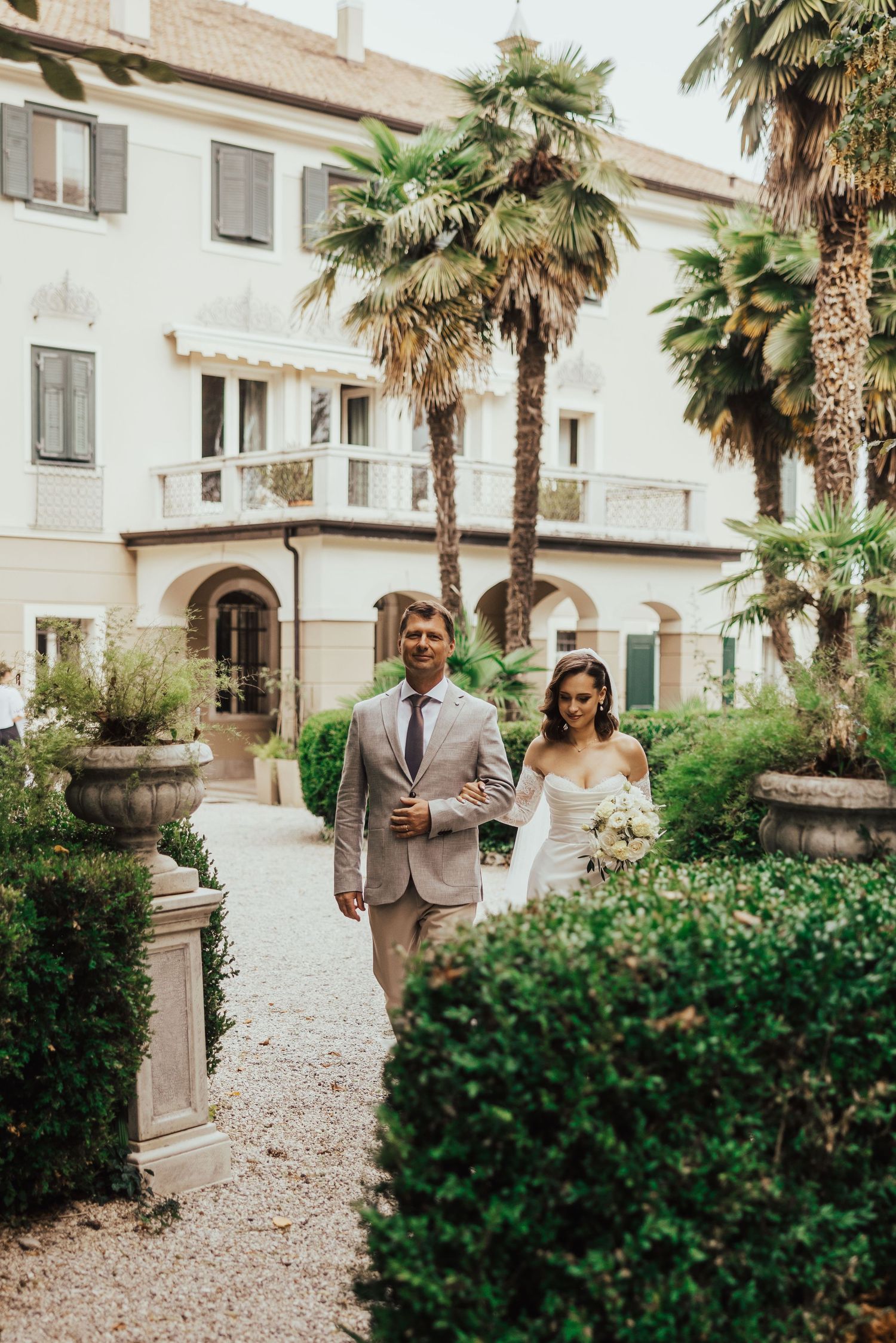 Bride and escort walking through Italian villa garden with palm trees, bride in white gown holding bouquet.