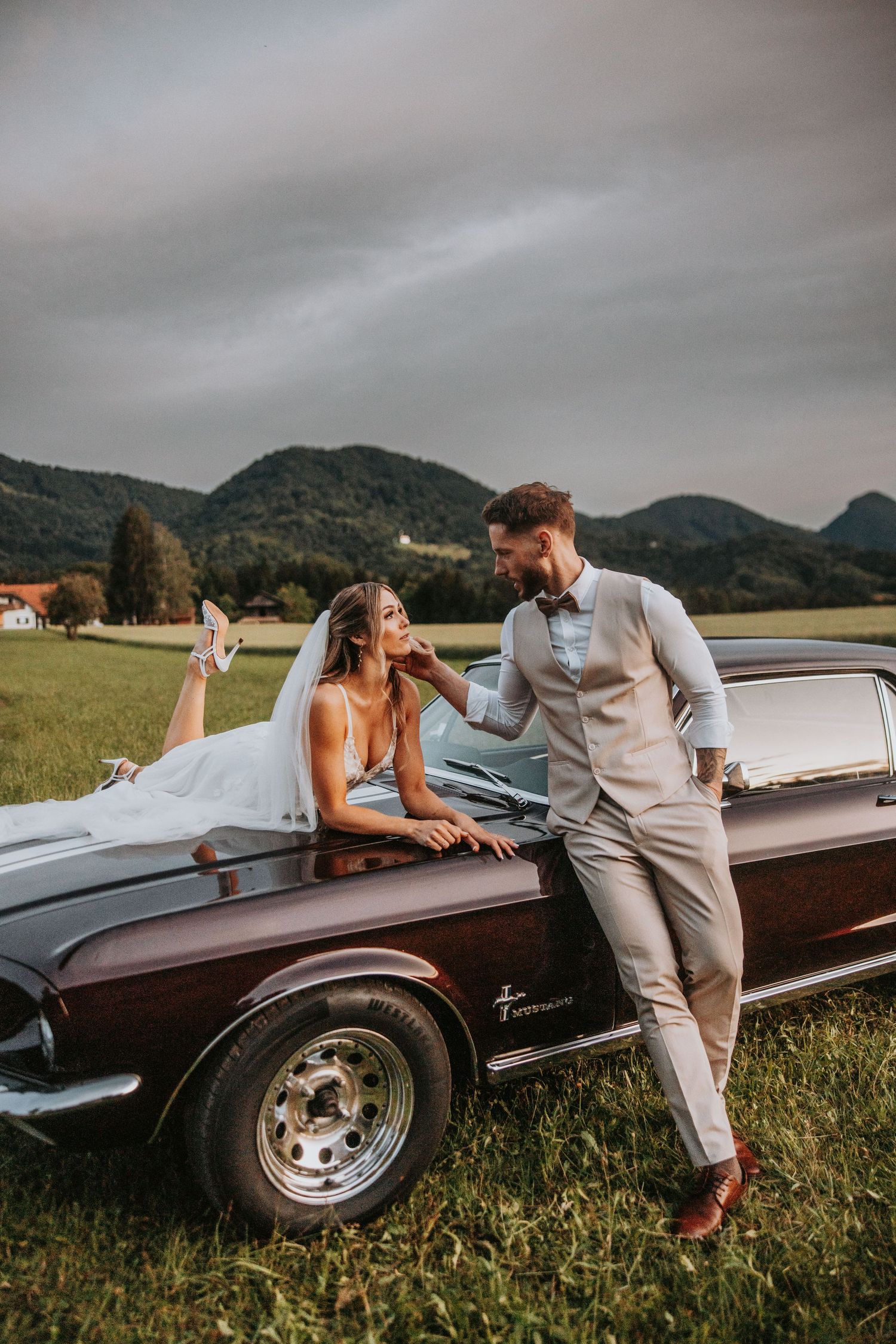 Bride and groom share romantic moment leaning on classic vintage car in scenic mountain meadow.