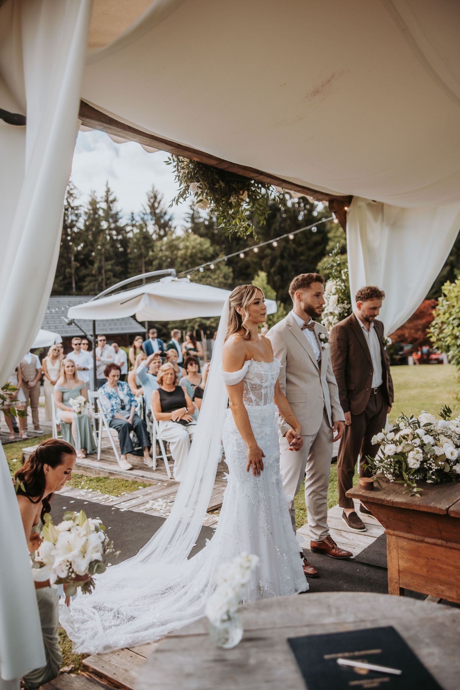 Bride in lace gown and groom in beige suit stand at altar during outdoor garden wedding ceremony.