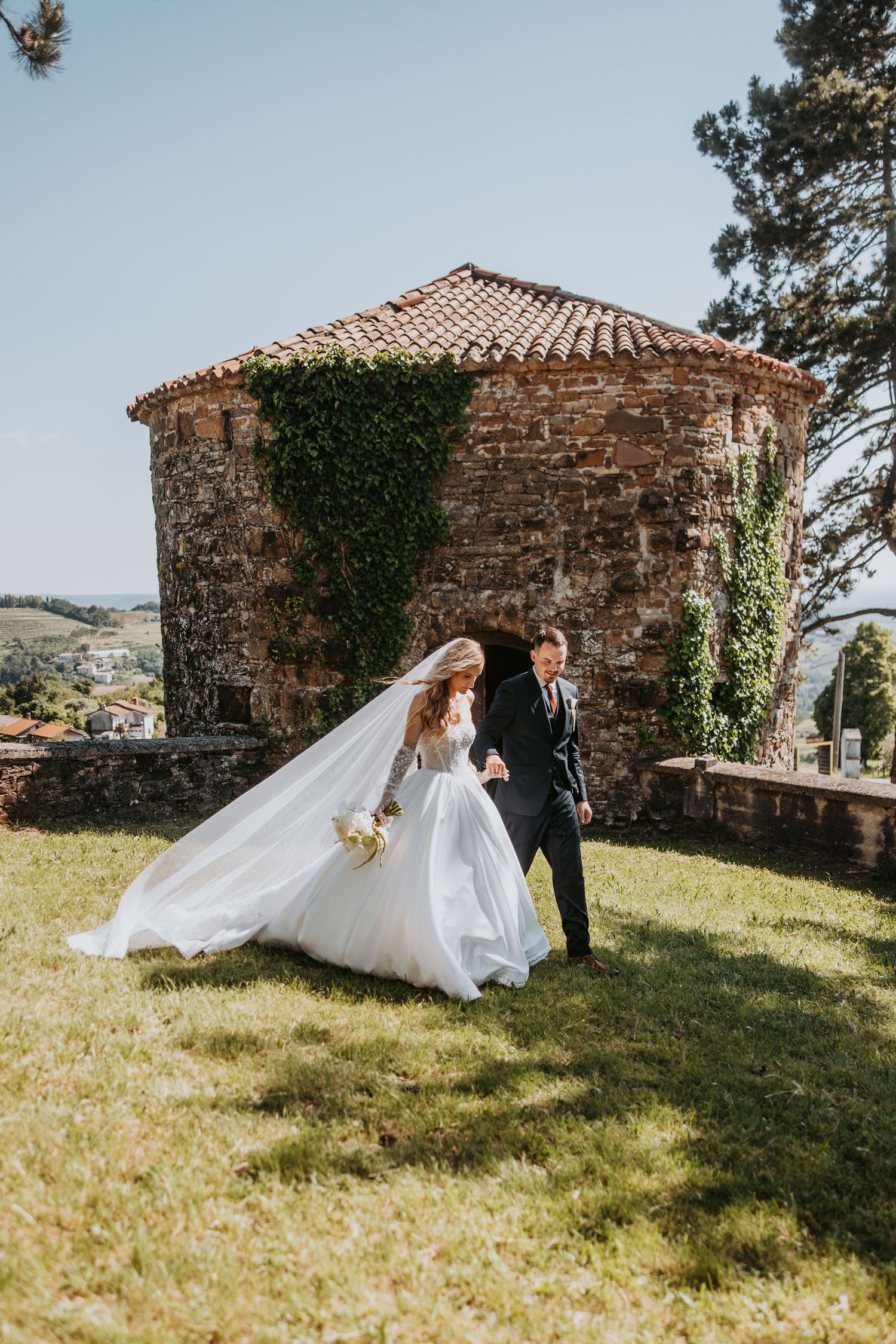 Bride and groom walking hand in hand on grass beside a stone ivy-covered chapel on sunny day.