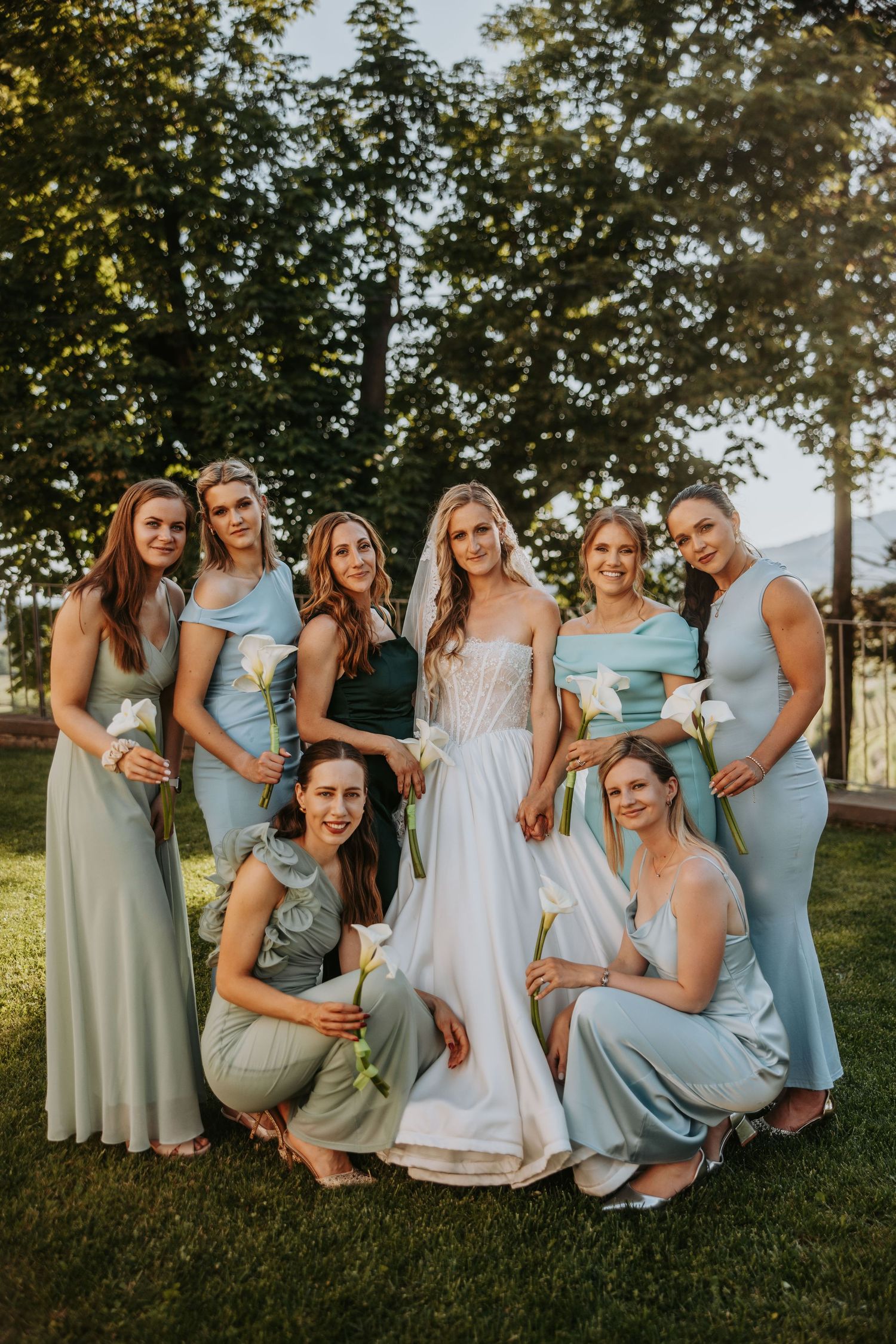 Bride in white corset gown poses with seven bridesmaids in sage and light blue dresses on green lawn.