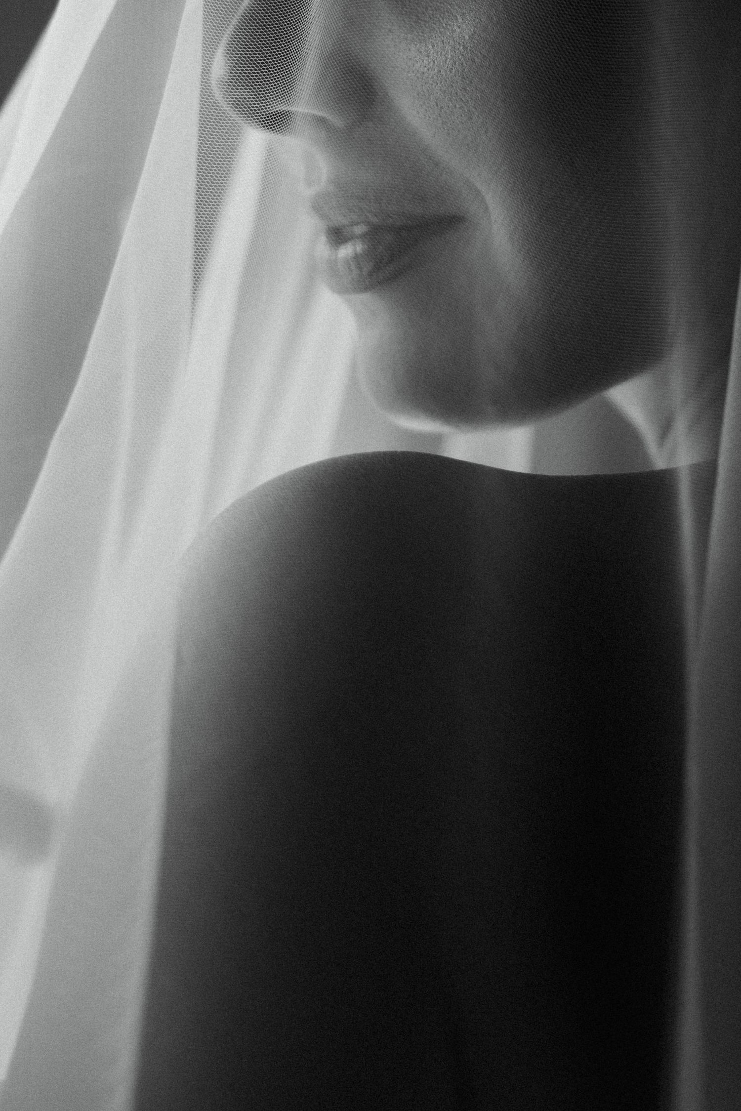 Black and white portrait of a bride wearing a veil, soft light highlighting her bare shoulder and gentle smile.