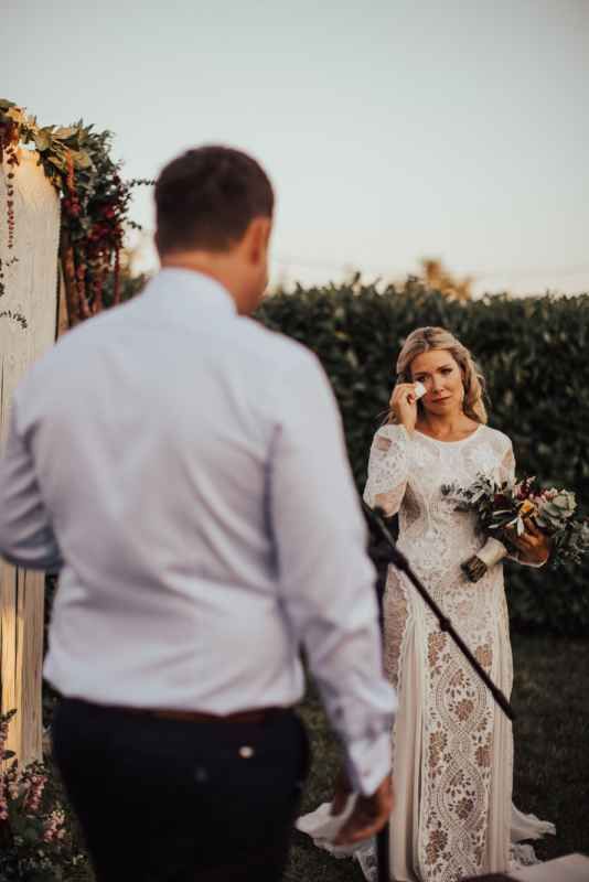 Emotional bride in lace wedding dress wiping tears holding bouquet during outdoor ceremony at sunset.